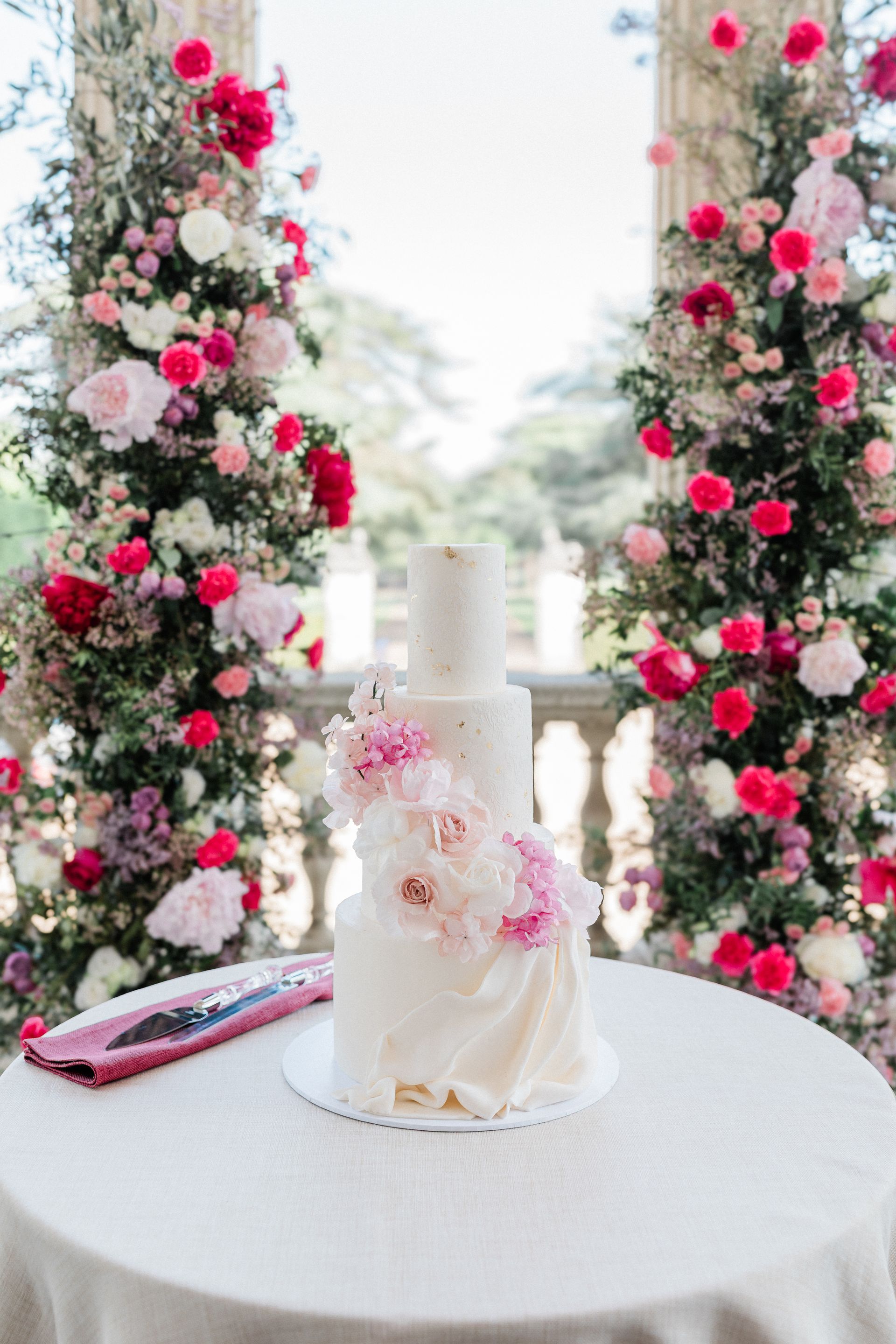 3-tier wedding cake with pink sugar flowers displayed in front of the wedding ceremony flower arch