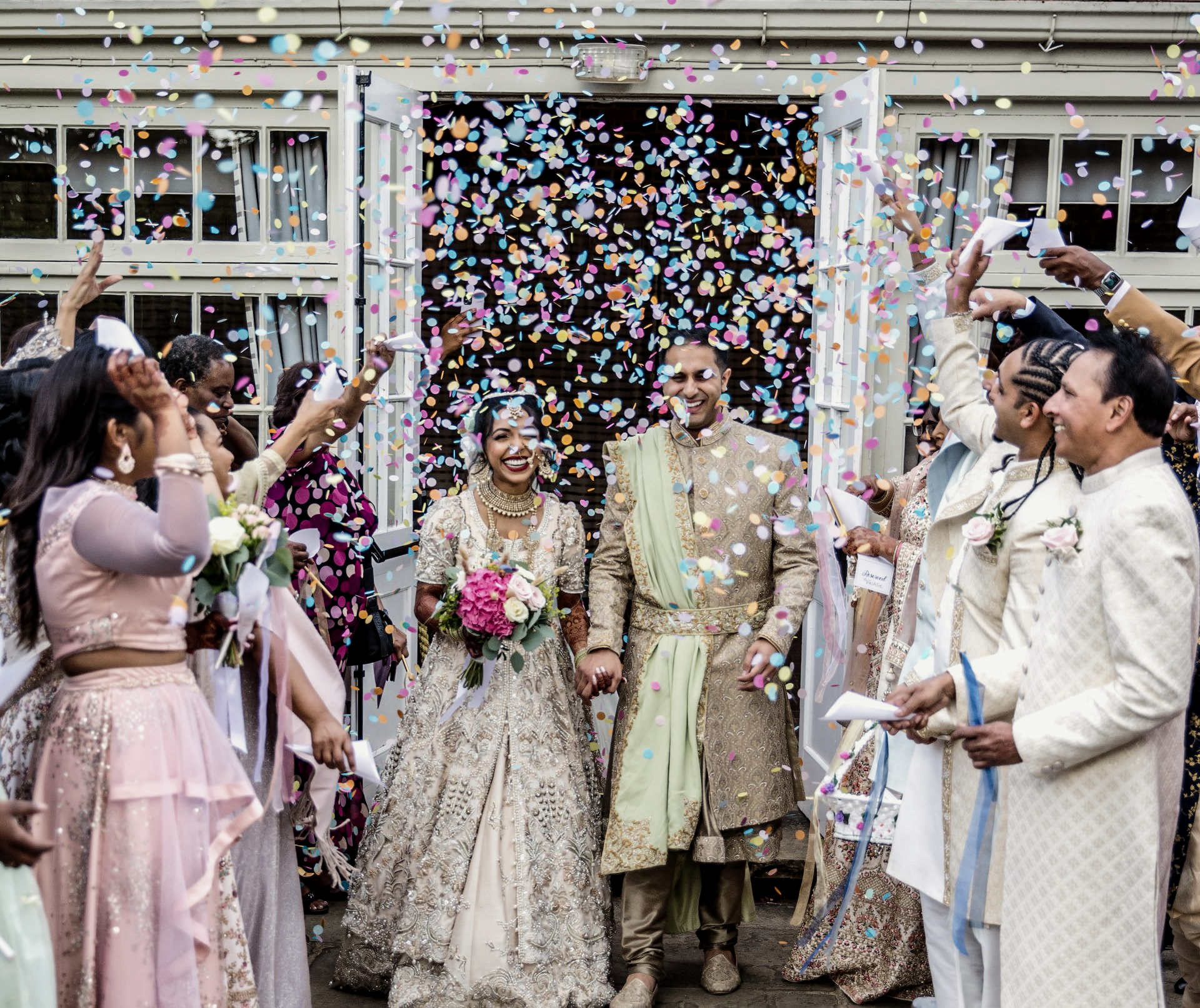 An Asian bride and groom smile as they exit a dry-hire wedding venue in South East England, showered in colourful confetti by guests, enjoying the present moment through professional coordination by Natasha Grant