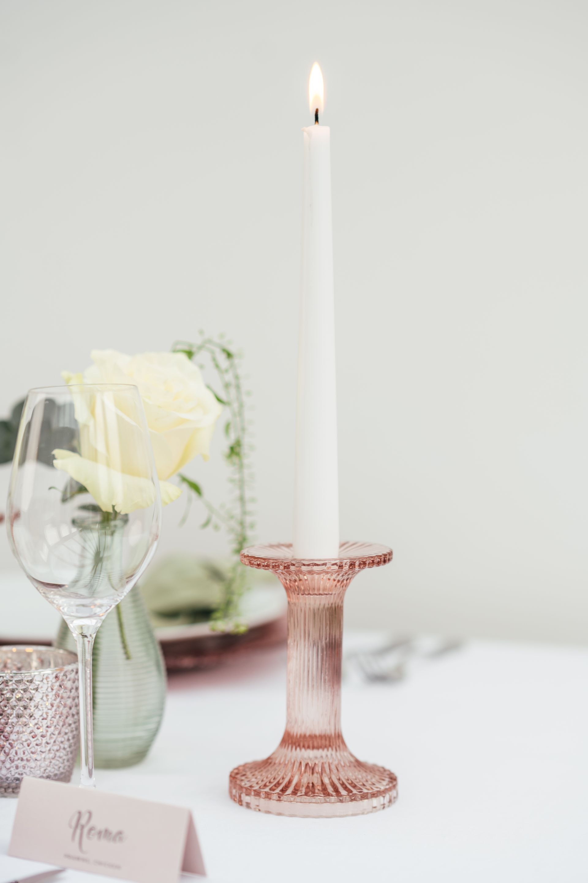 Pink glass candlestick with white candle displayed on a table with pink tea light and cream rose in bud vase