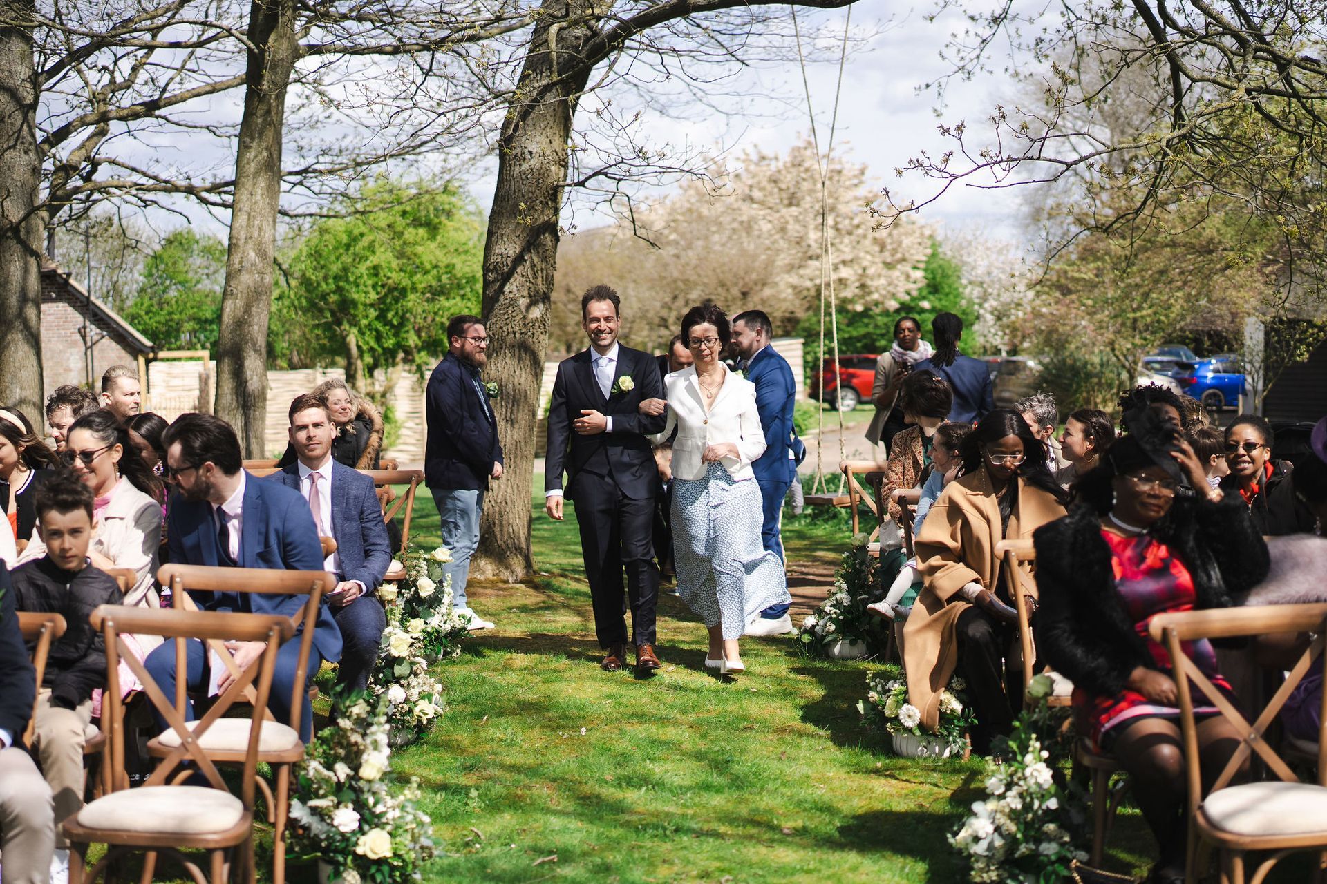 White groom male bin a blue suit walking down the aisle outside in a field with his Mum