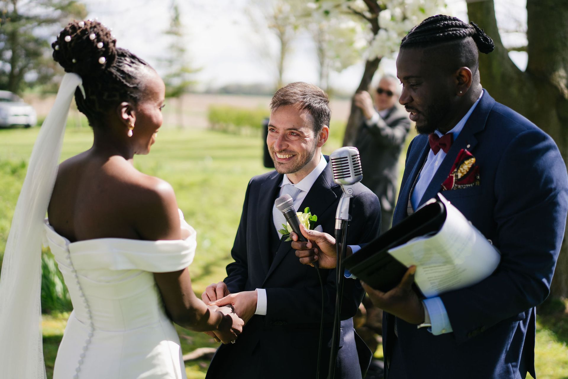 Black bride in a white dress holding hands and smiling at white male groom in his blue suit during their outdoor wedding ceremony