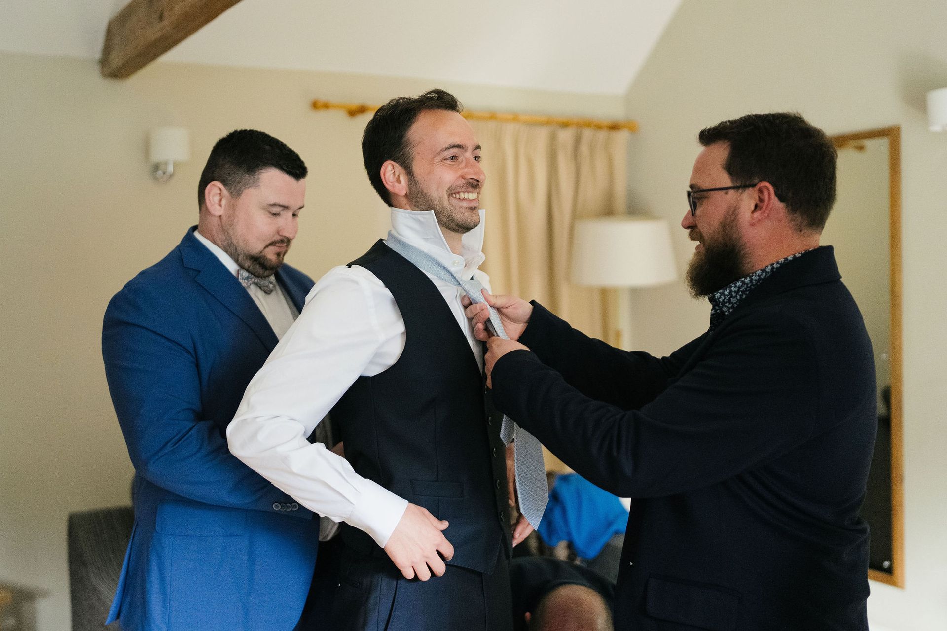 White groom in a blue waistcoat having his tie put on by 2 groomsmen