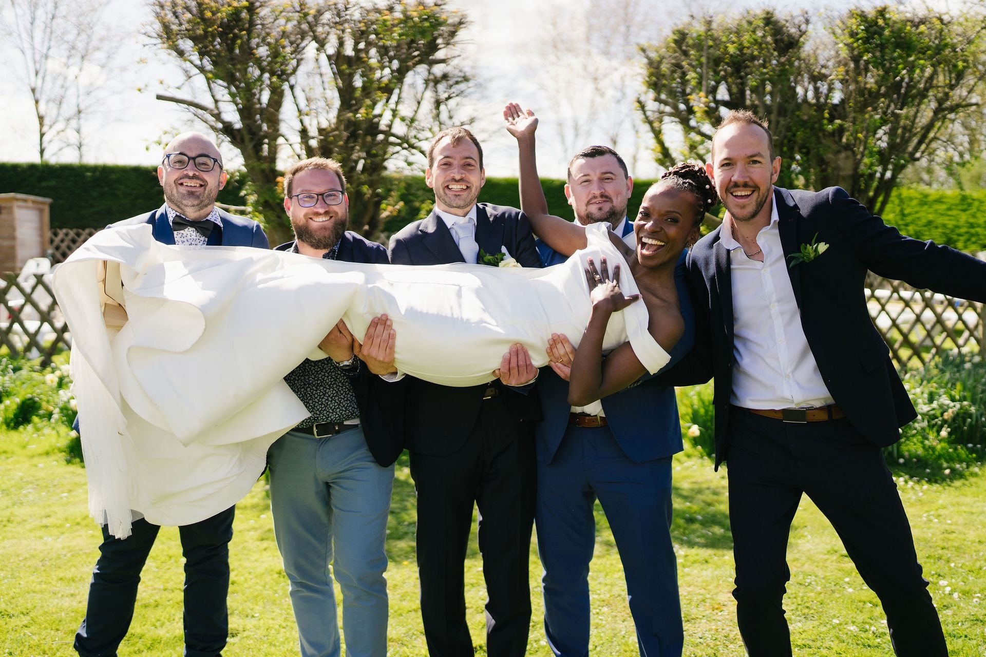 Bride in white dress been held horizontally by 5 groomsmen outside