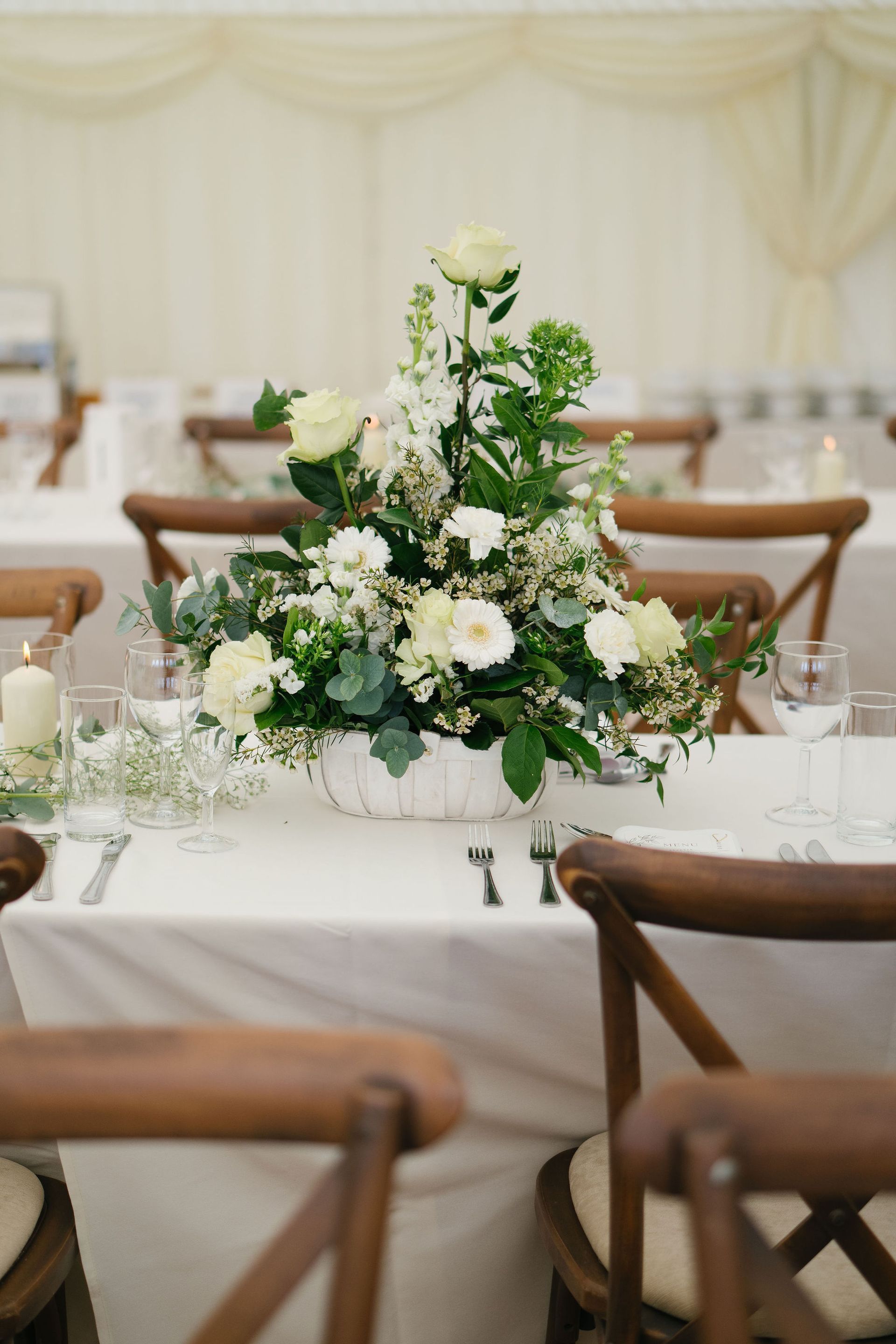 White flowers with green leaves displayed on a long wedding reception table with brown cross back chairs