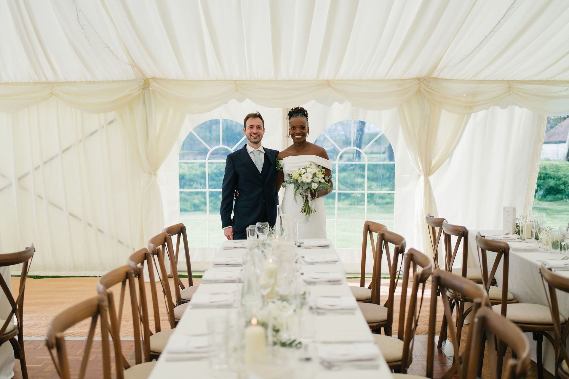 Bride and groom standing at the head of their long wedding reception table in a white marquee smiling at the camera