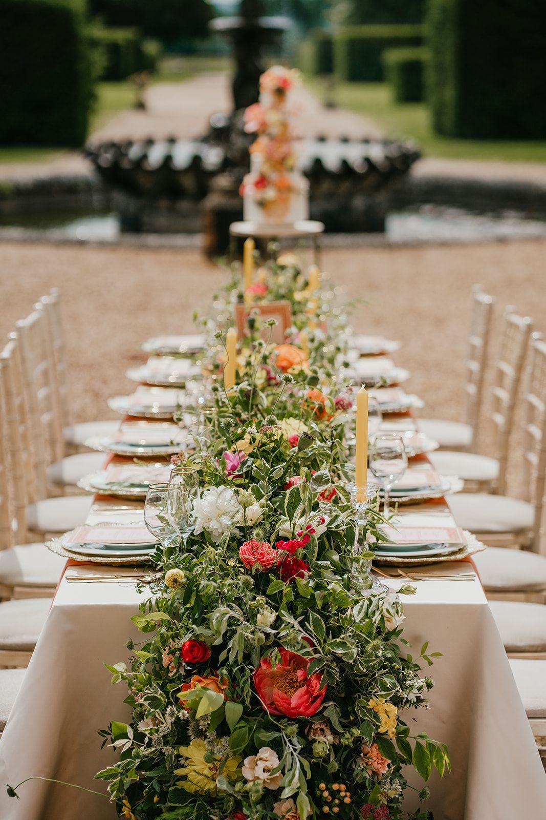 Wedding table set up outside by a fountain with colourful floral runner and wedding cake