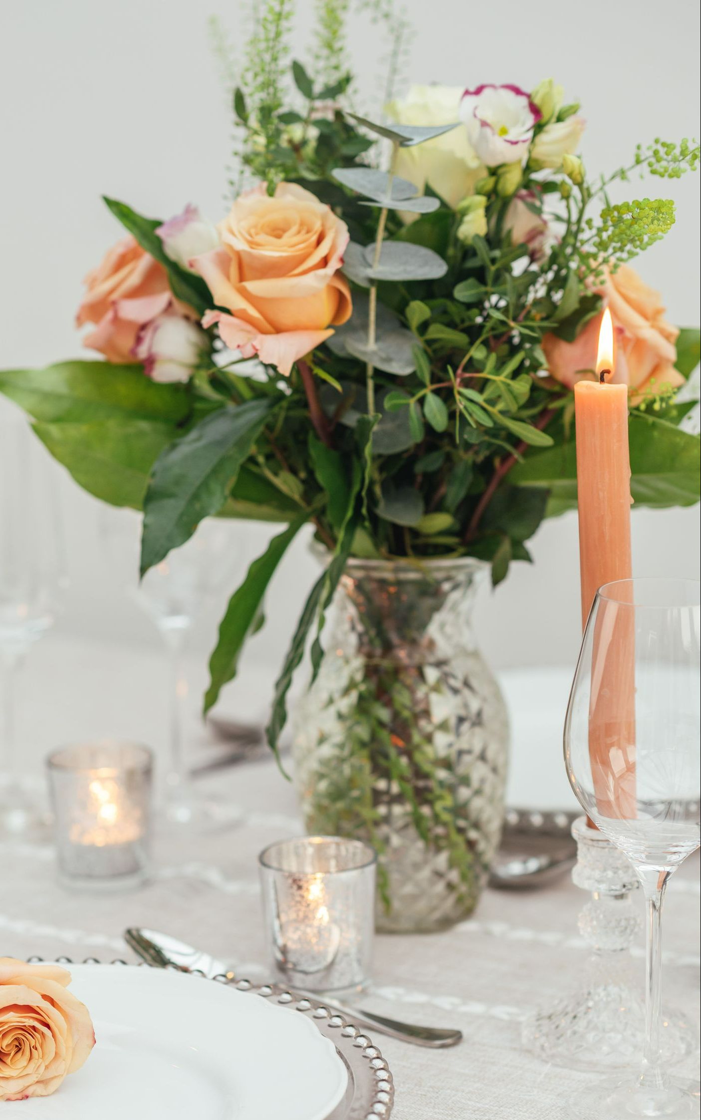 Clear textured vase holding orange flowers displayed on a table with silver tea lights and an orange candle