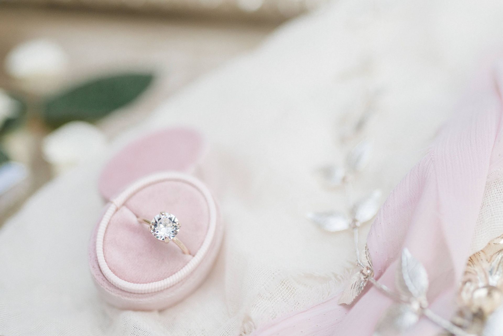 A diamond ring in a pink oval ring box displayed next to cream fabric