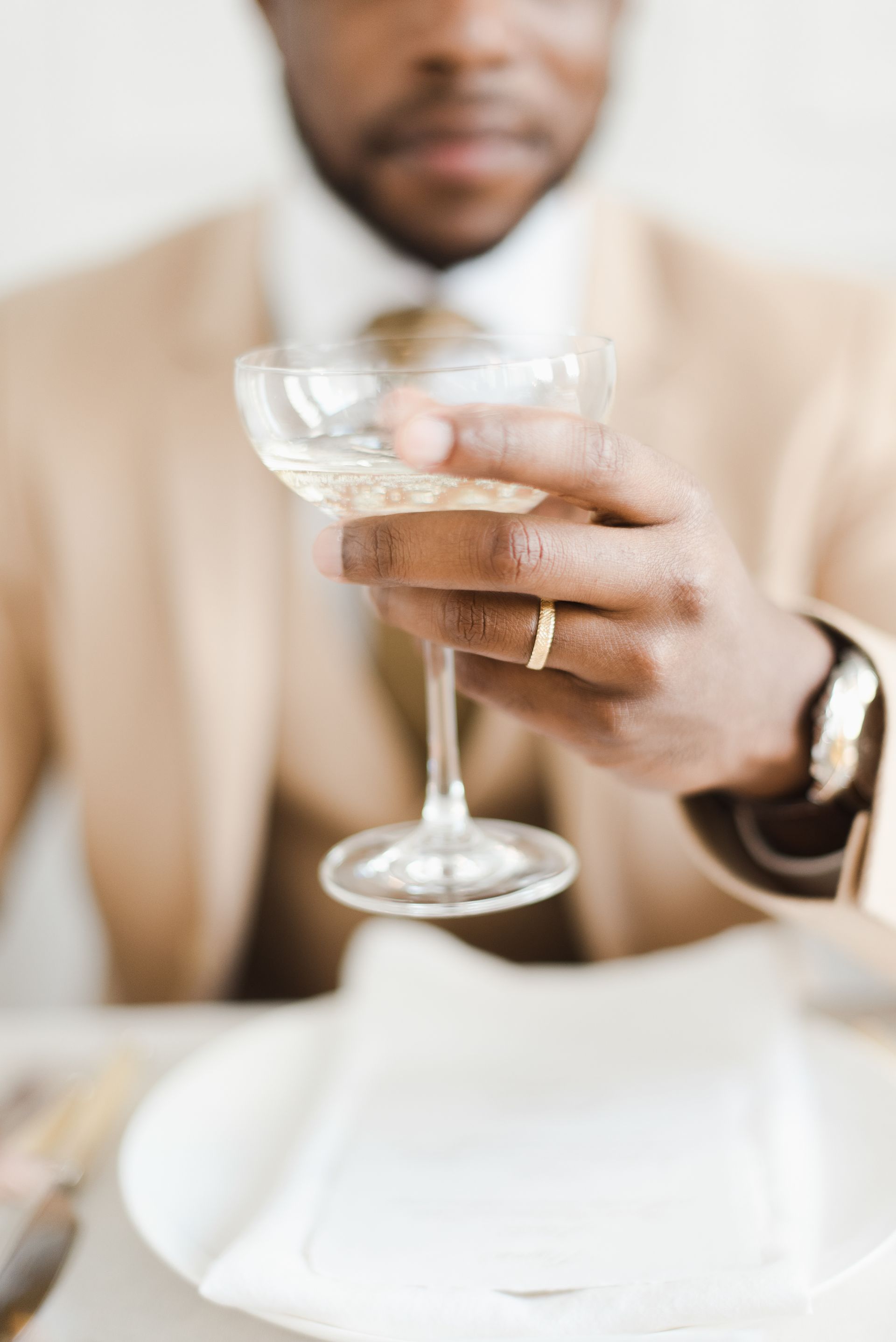 Black groom in light brown suit with gold ring holding up a glass of champagne to the camera
