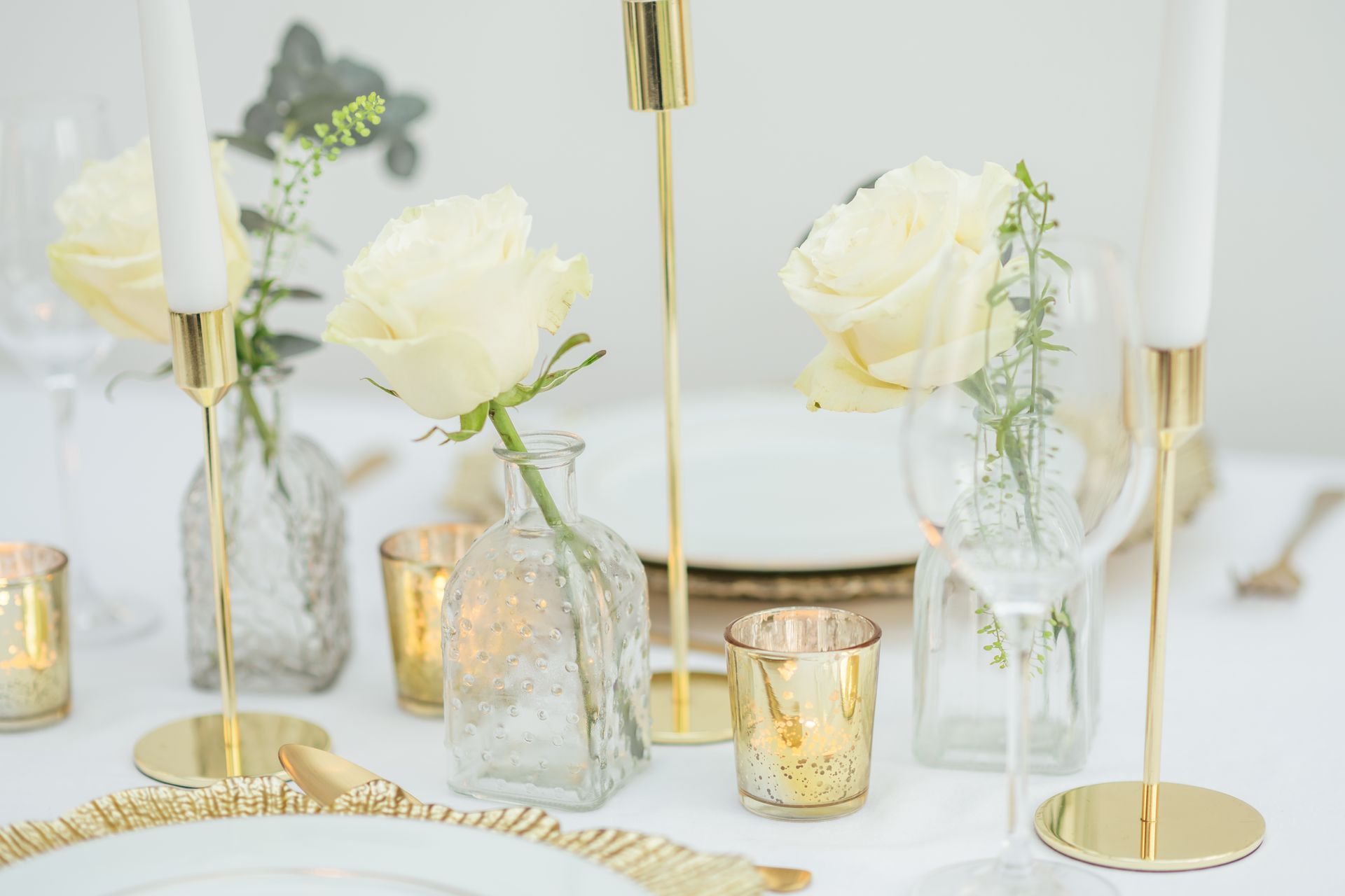 3 clear bud vases with cream roses in them displayed on a table with gold tea lights