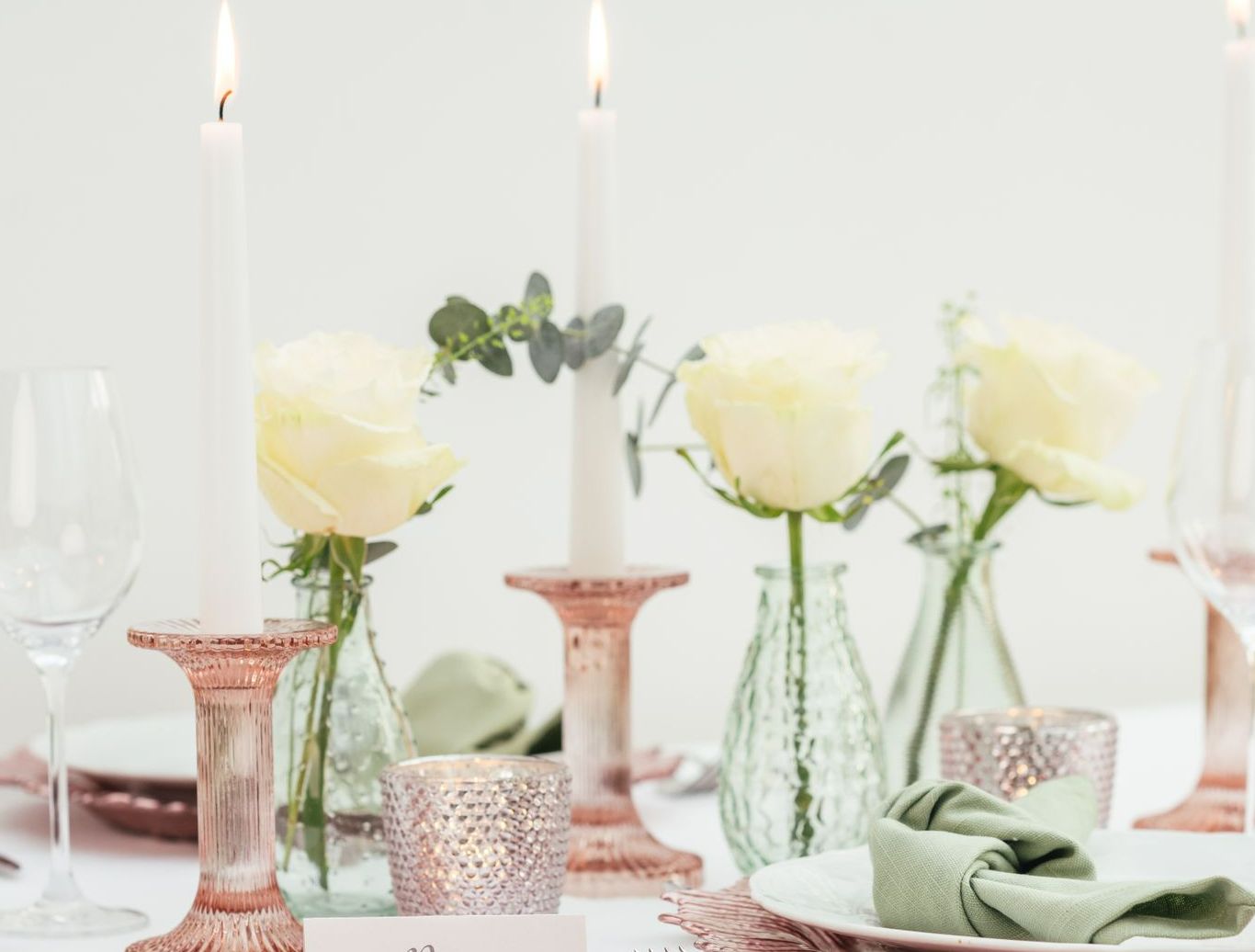 Pink glass candlesticks and white candles displayed on a table with cream roses in vases