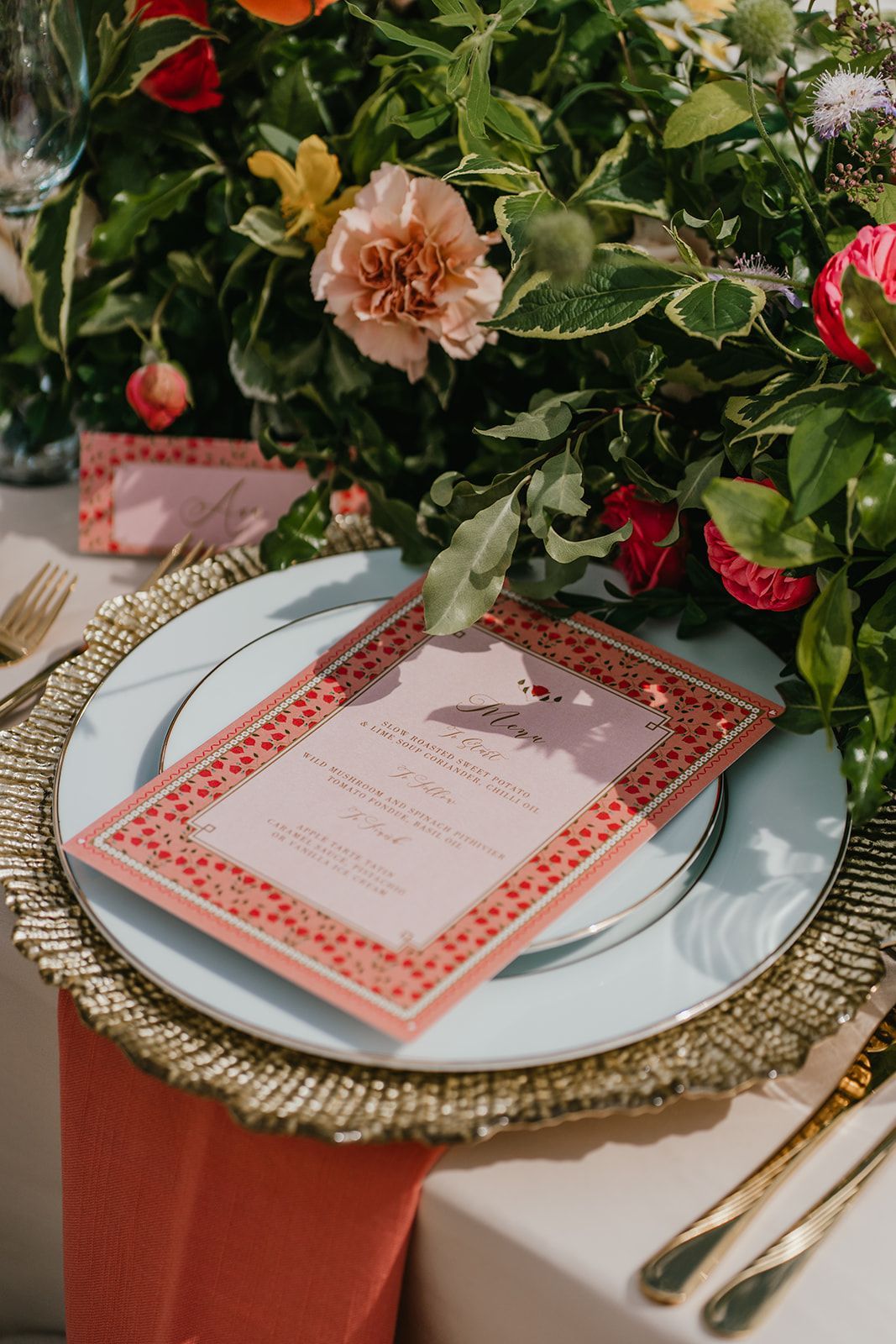 Close up of a white plan with gold edging with  a gold charger plate below it and an orange coloured menu on top of the plate, surrounded by orange and pink flowers and green foliage
