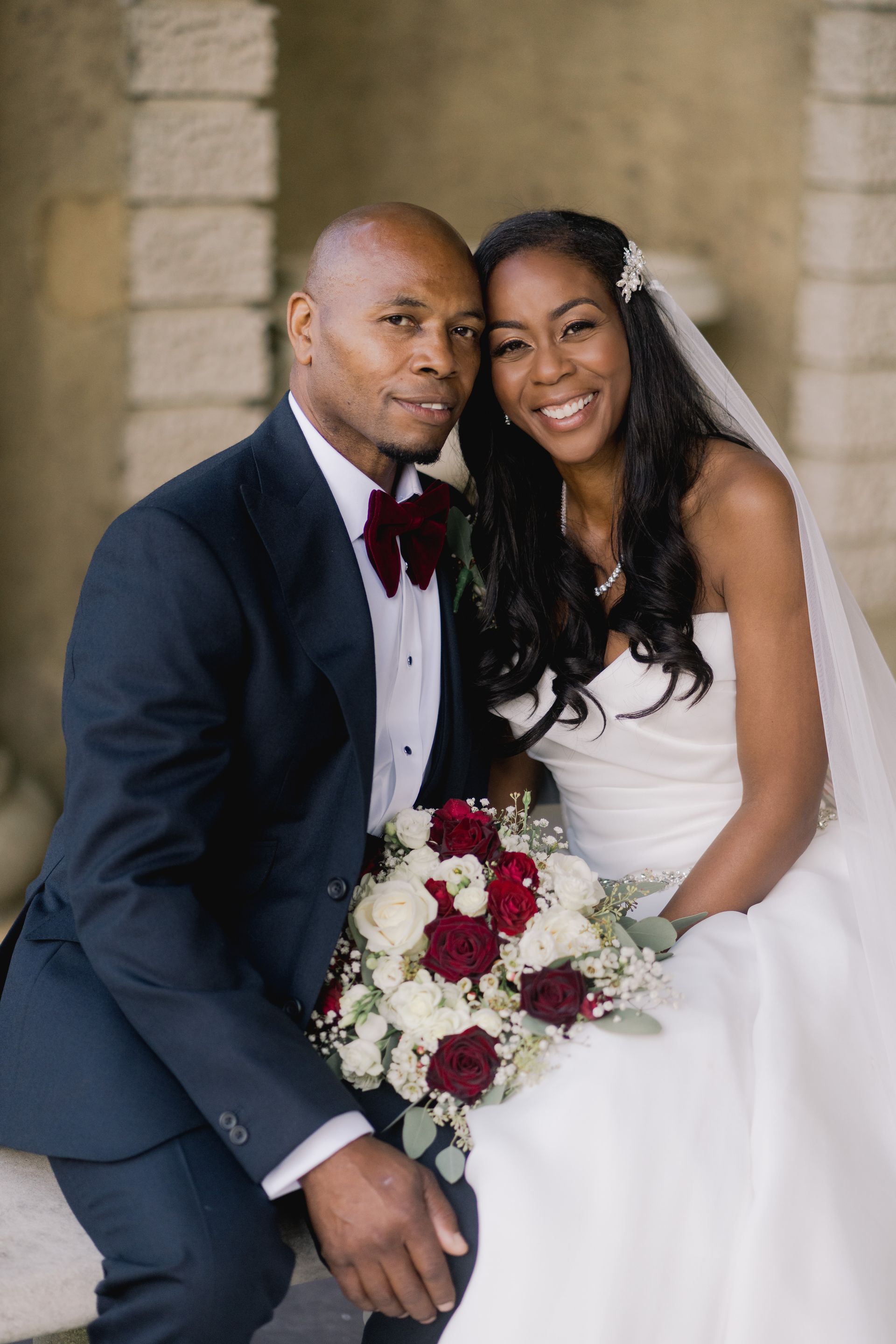 Black bride and groom smiling at the camera with the bride holding white and red floral bouquet