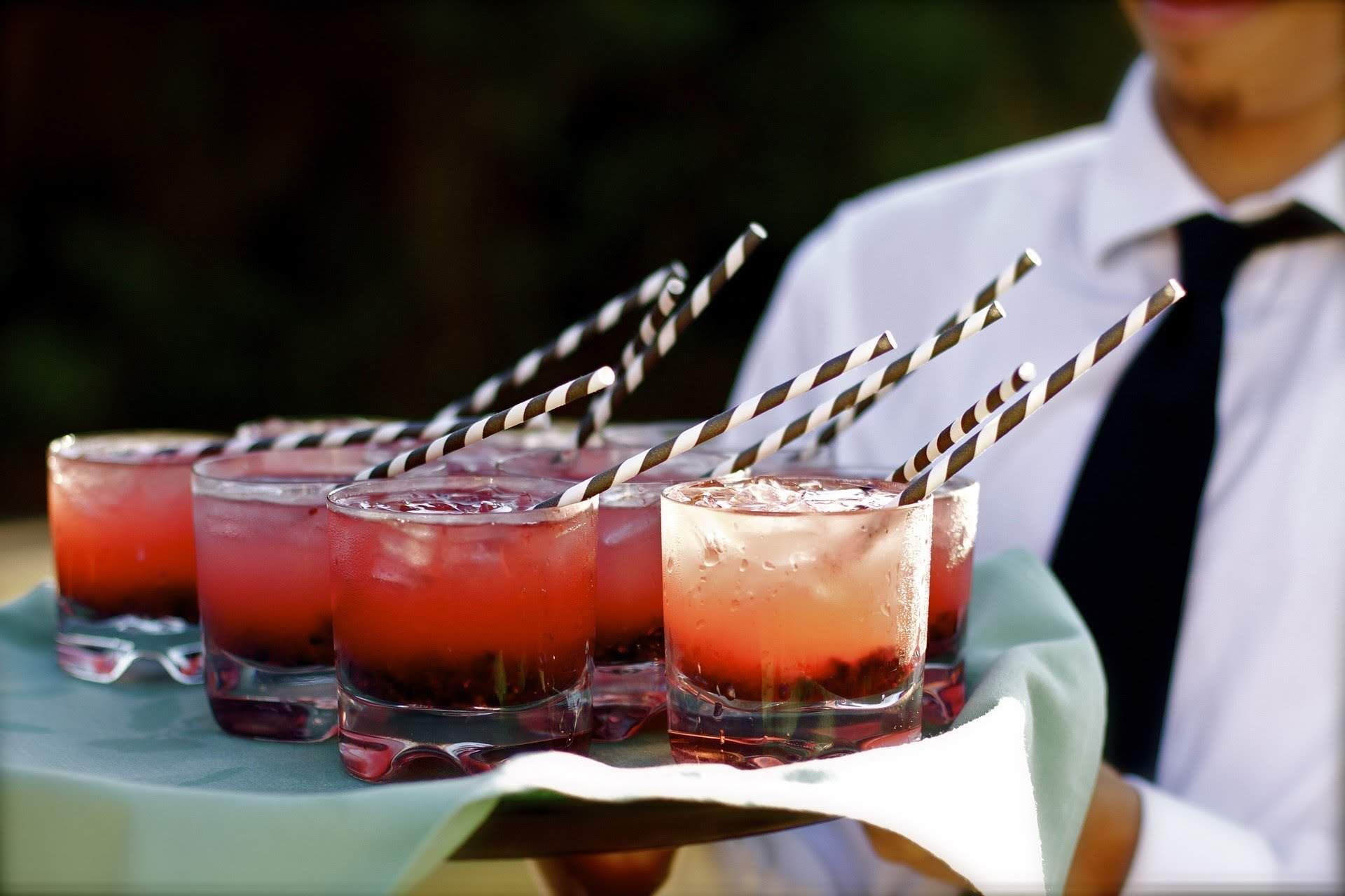 Bar man holding a tray of red cocktails with fruit in it and black and white stripy straws