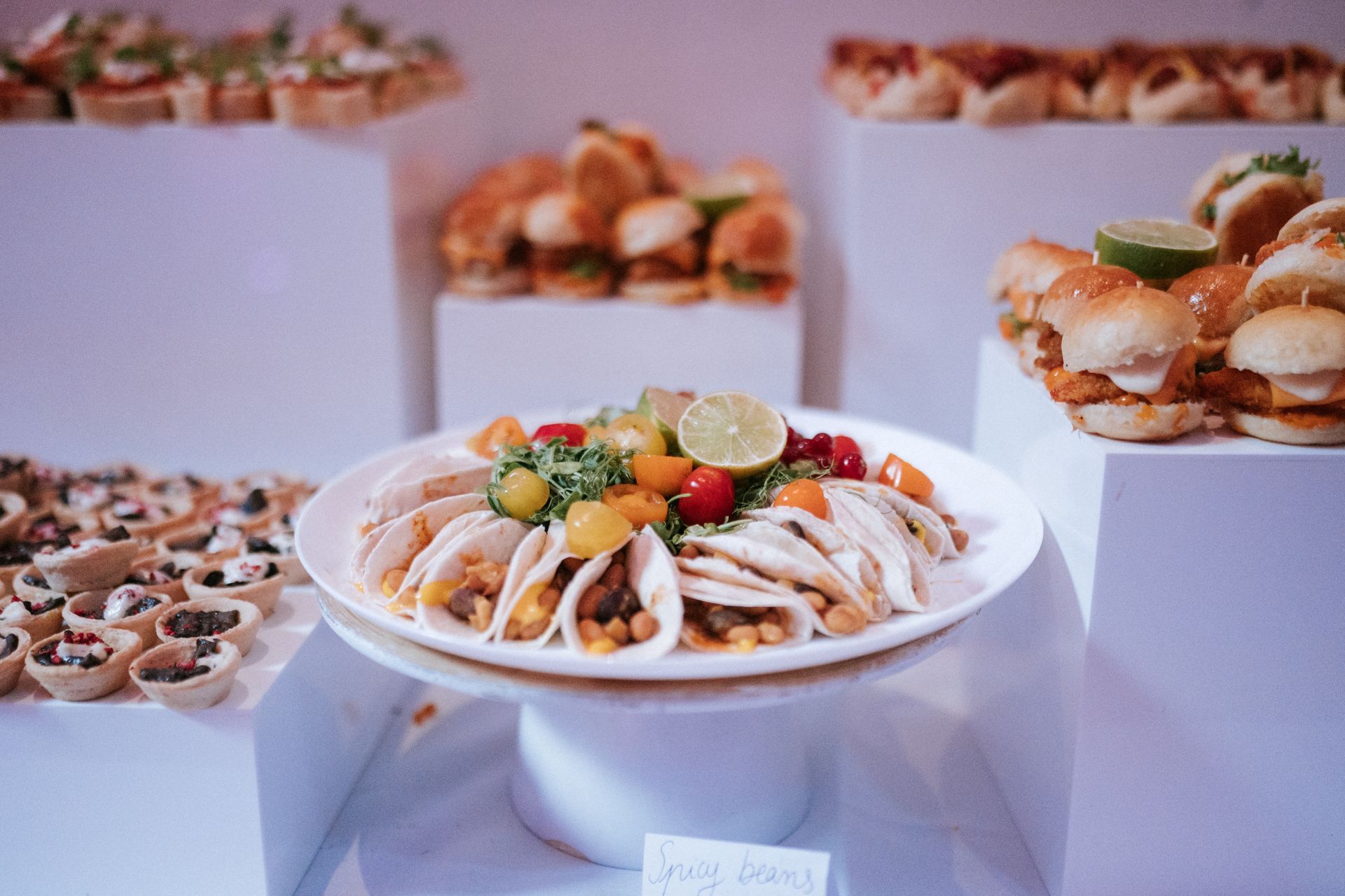 Picture of a buffet food table displaying spicy beans in wraps and mini chicken burgers on white stands