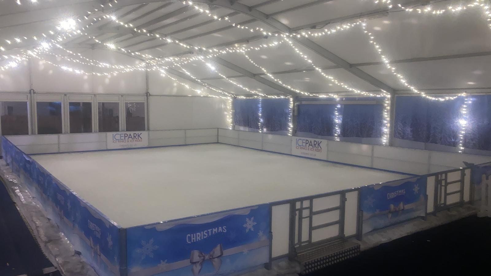 Small beautiful ice rink decorated with lots of white lights over and blue Christmas vinyl's on the barriers