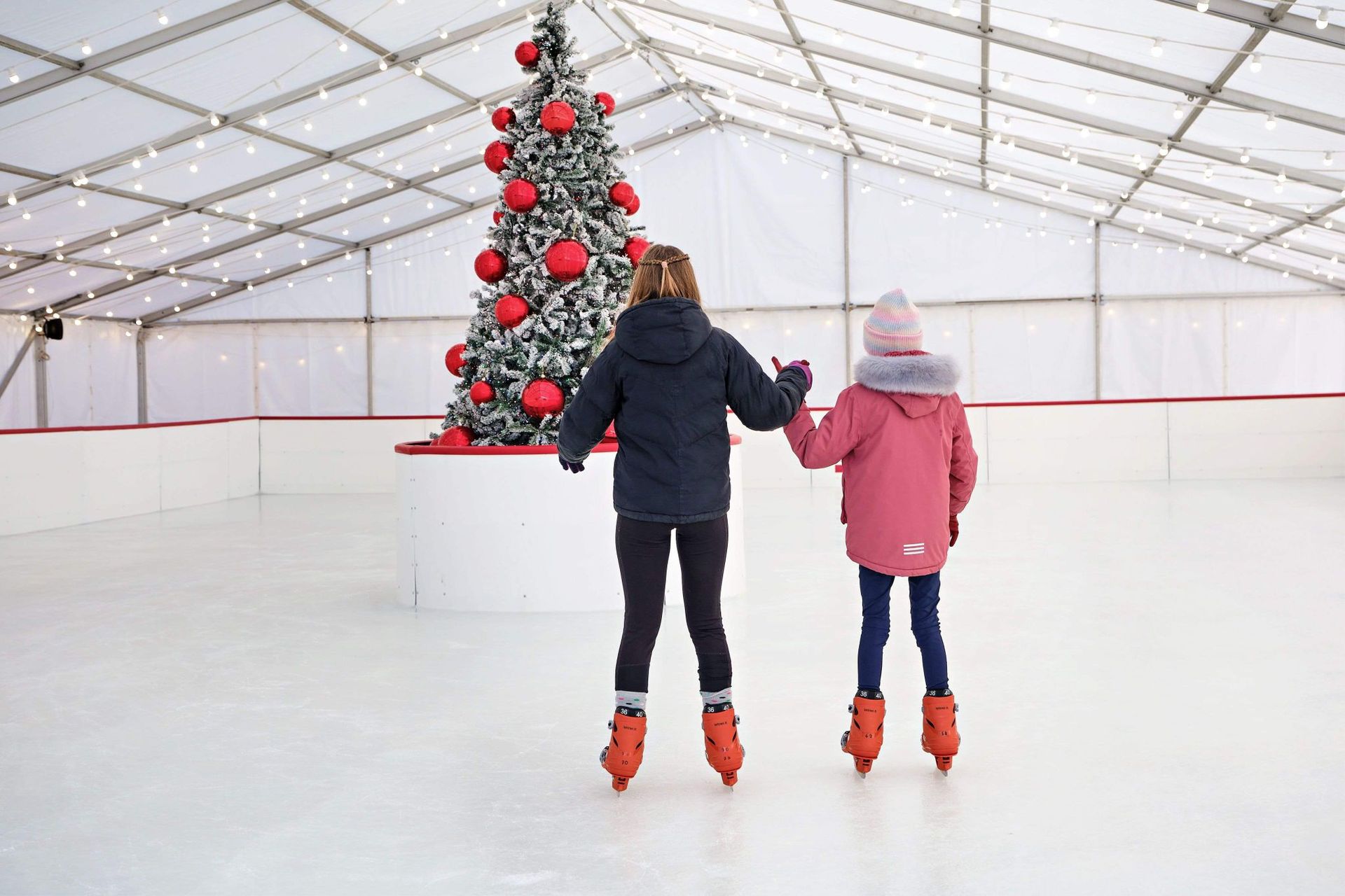 Two children ice skating with a beautifully decorated Christmas tree in the centre of the ice rink.