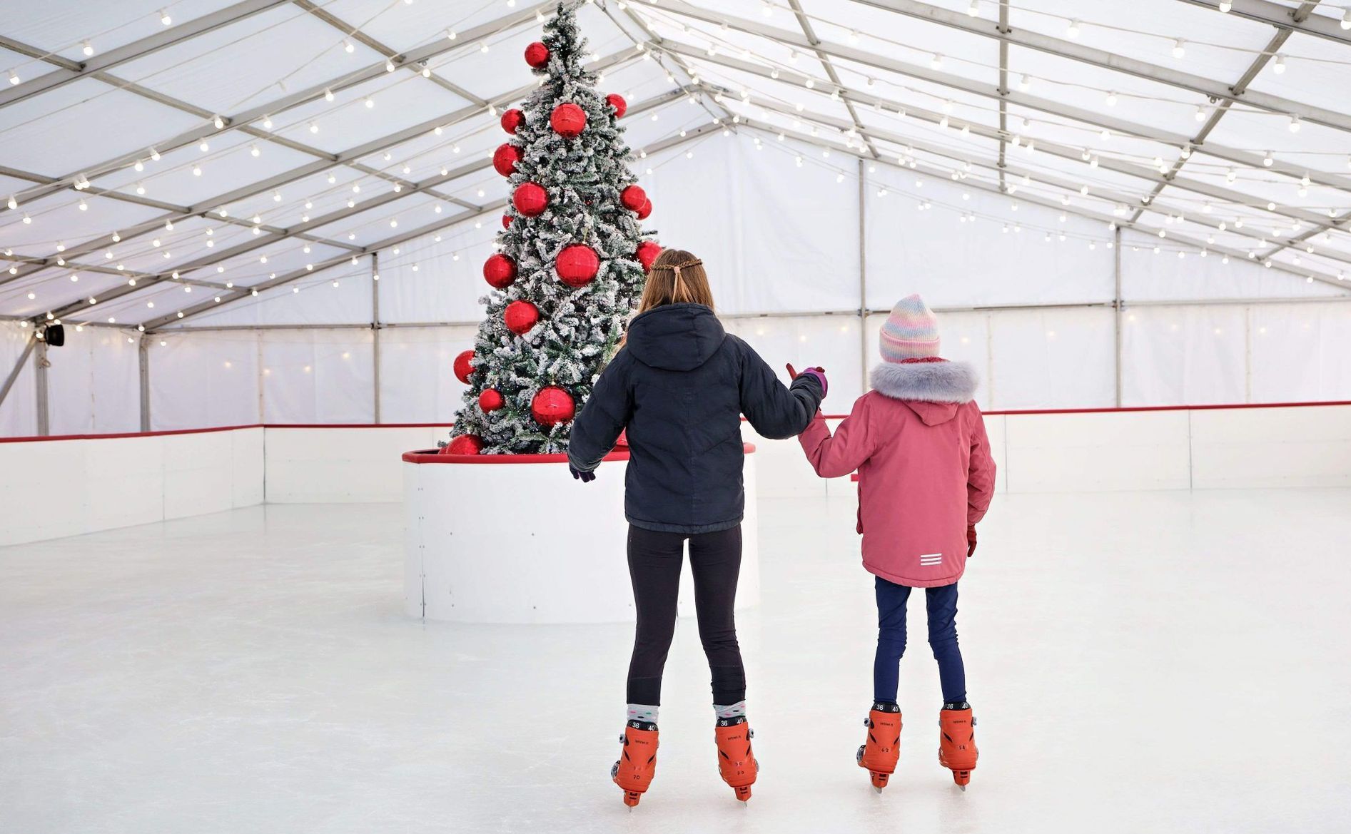 Two young girls holding hands wearing winter coats ice skating.  Christmas tree with bright red baubles.