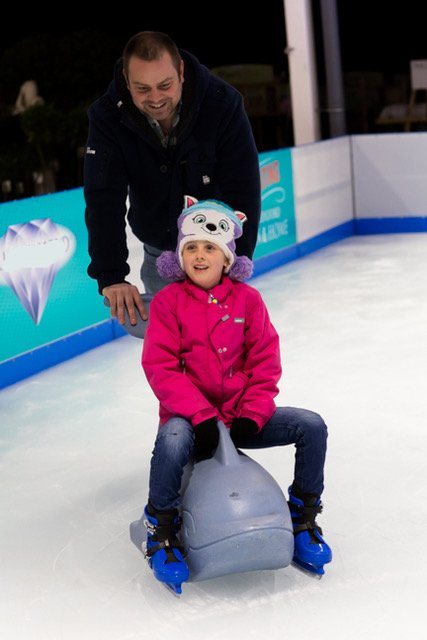 Dad ice skating and smiling. Daughter with a wide smile on a dolphin skate aid.