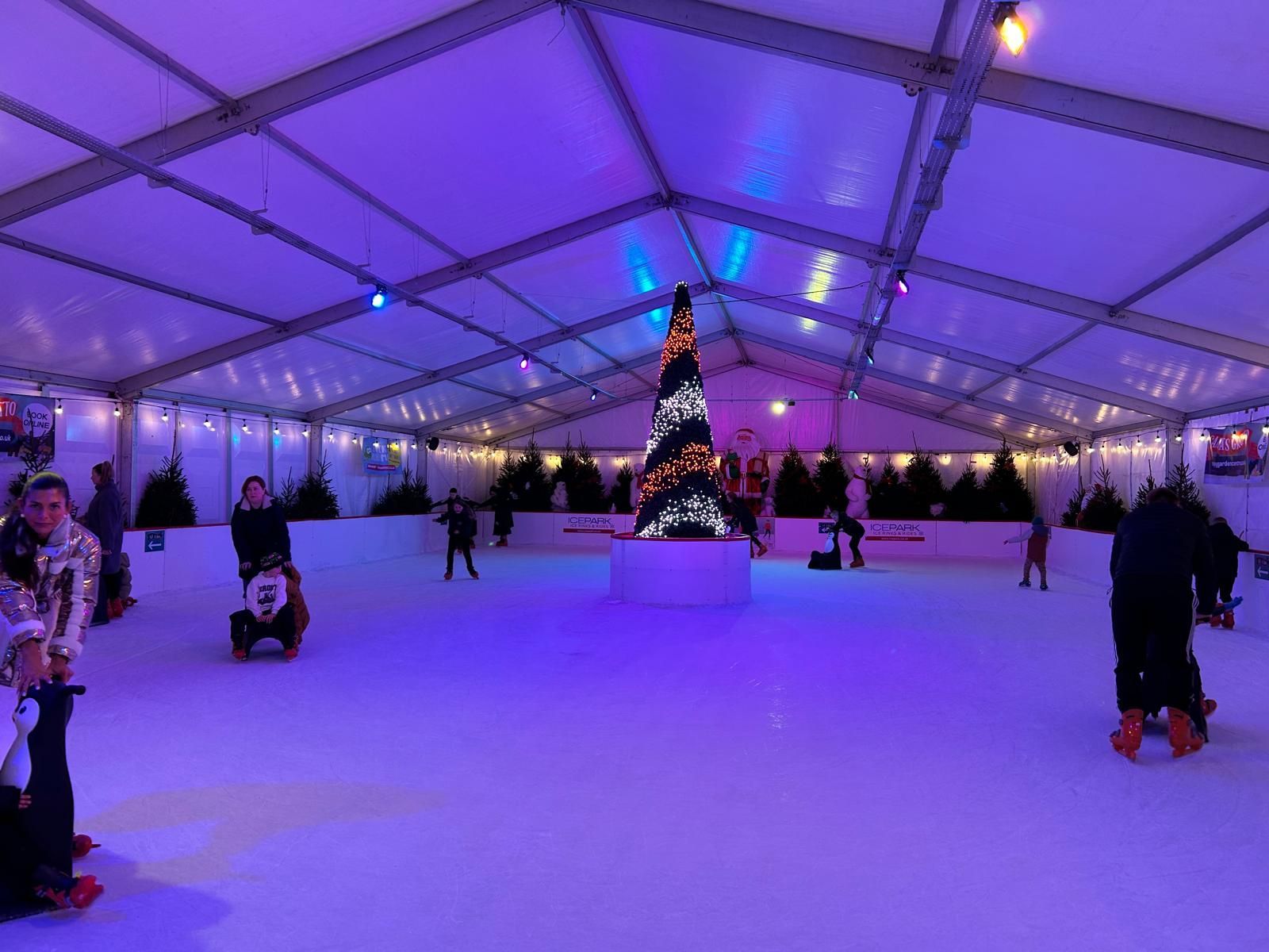 Ice rink lit up bright purple with lots of skaters and decorated Christmas tree in the centre