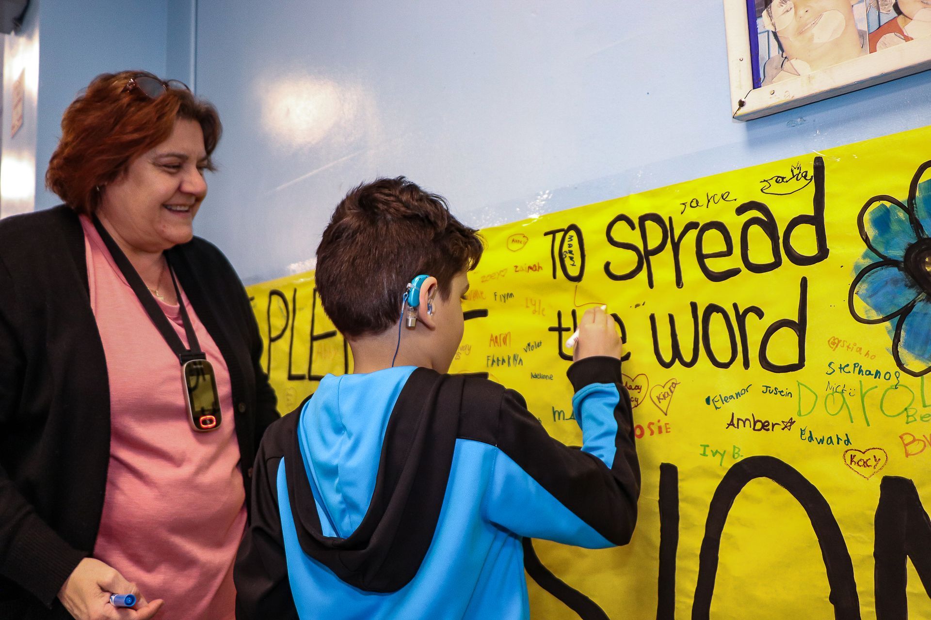 A young boy with a hearing aid writes on a banner about inclusion. A woman in pink is smiling as the boy writes.