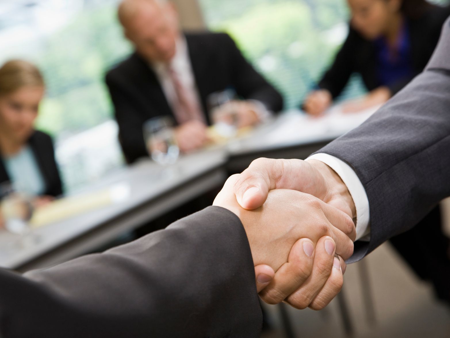 Close up of handshake with blurred image of business meeting in the background.