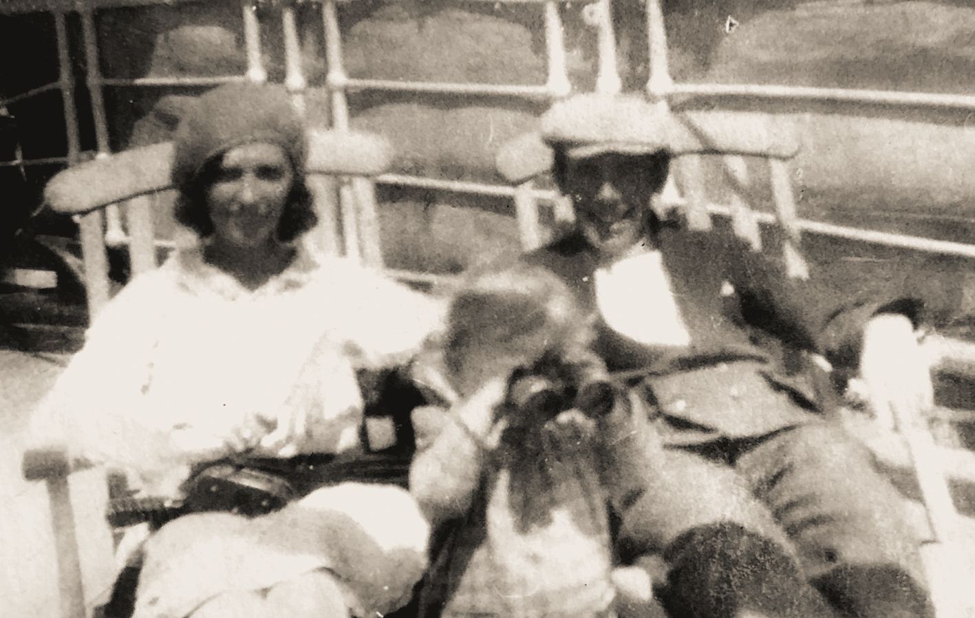 Schwarzweiß-Fotografie aus dem frühen 20. Jahrhundert: Drei Personen sitzen auf Holzliegestühlen auf dem Deck eines Schiffes. Links eine Frau mit hellem Kleid und Hut, rechts ein Mann mit dunkler Kleidung und Mütze, in der Mitte ein Kind mit Fernglas vor dem Gesicht. Im Hintergrund sind Reling und Schiffselemente zu erkennen.Die Szene erzählt von Bewegung, von Übergängen und von der stillen Nähe zwischen Generationen auf dem Weg ins Ungewisse.