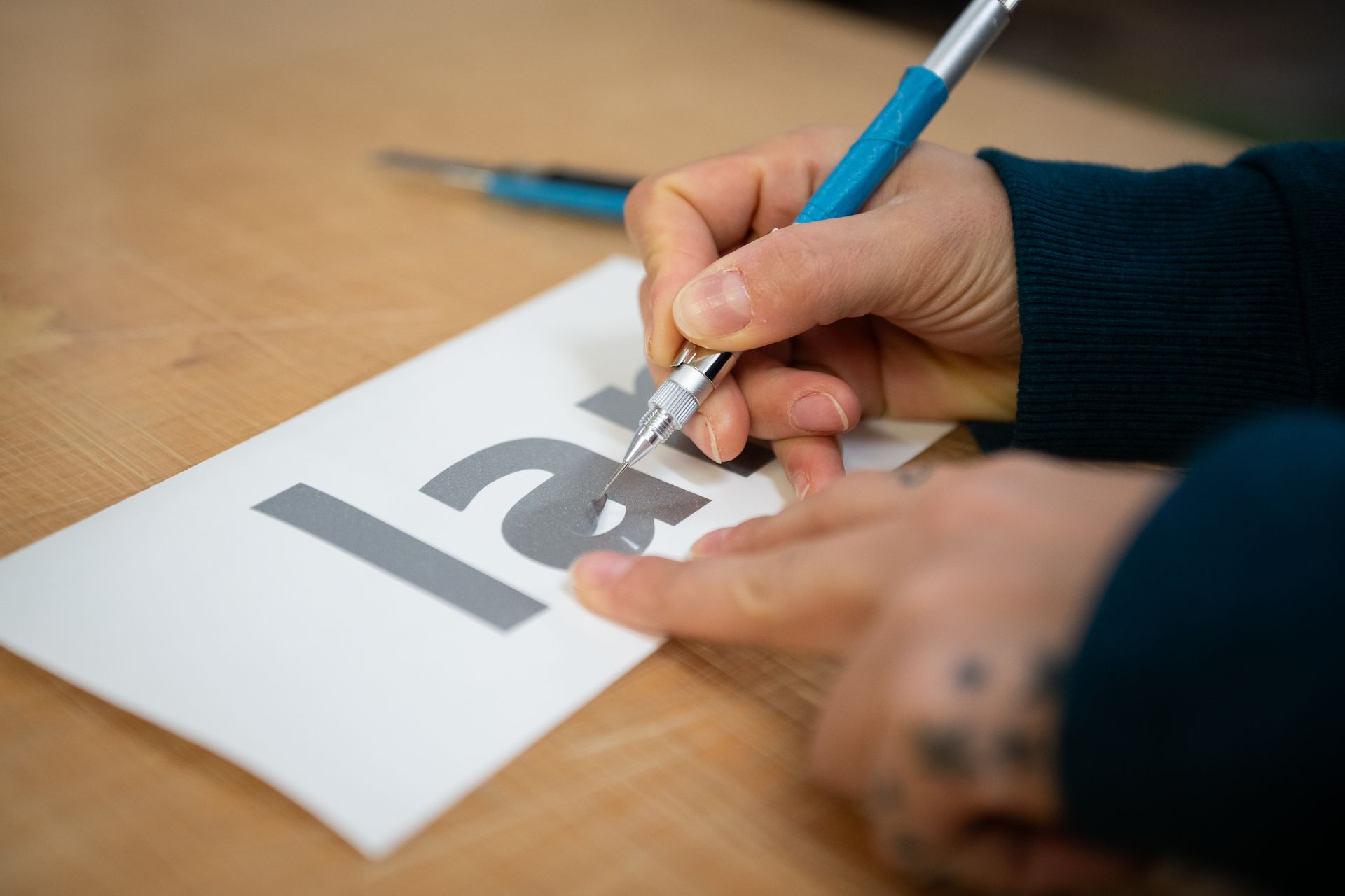Man sieht zwei Hände einer Mitarbeiterin in Nahaufnahme, die gerade dabei ist Buchstaben nach dem Plotten auf Klebefolie mit einem Werkzeug (ähnlich einer großen Nadel) auszulösen.