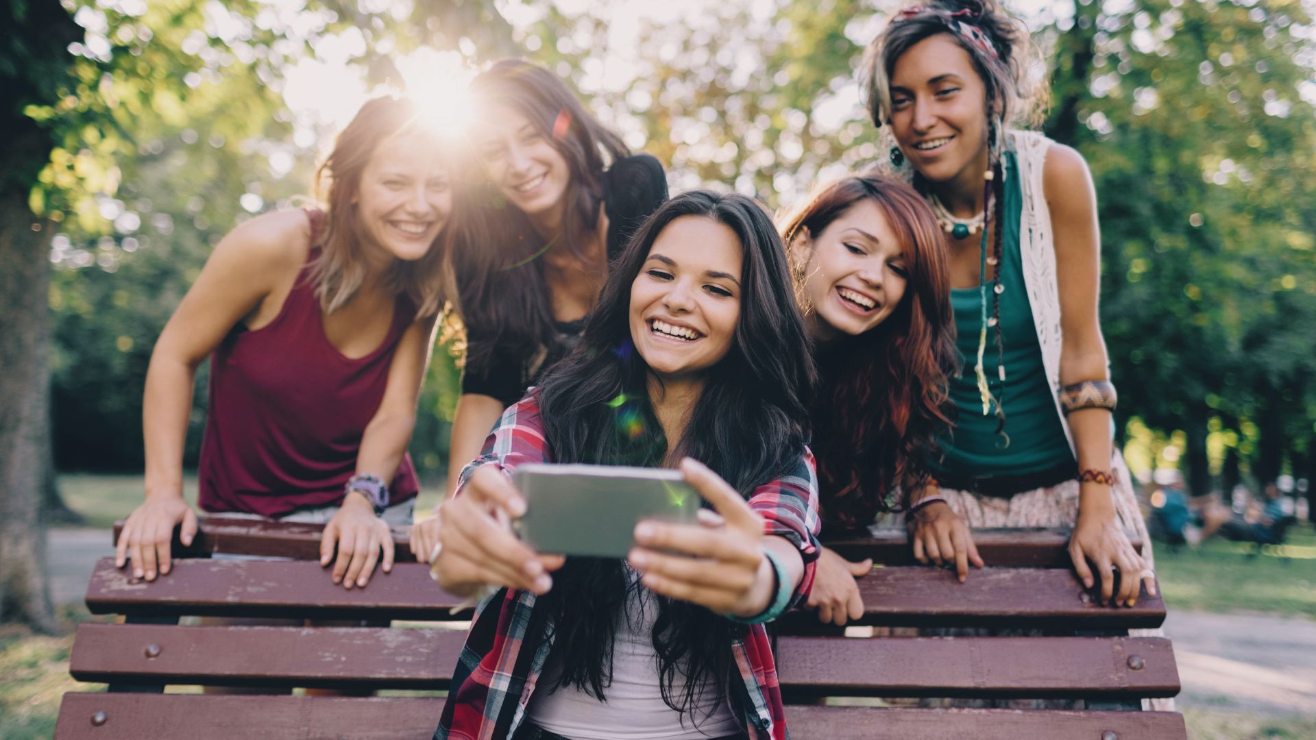 Gruppo di cinque amiche sorridenti che scattano un selfie su una panchina in un parco.