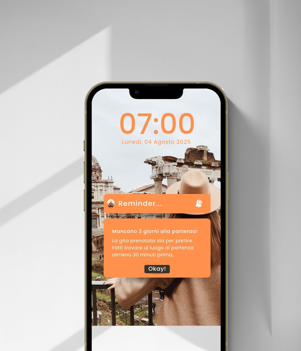 Smartphone con schermata di blocco che mostra un promemoria per un viaggio prenotato, con sfondo di una turista che osserva rovine storiche.