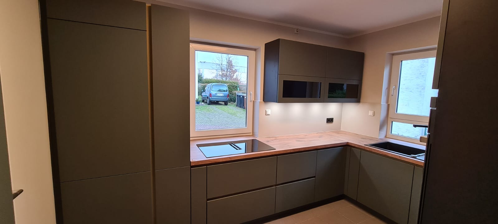 Das Bild präsentiert eine erstklassige Nobilia Küche mit grifflosen Schränken, die mit den exquisiten EASYTOUCH 964 Mineralgrün ultramatten Fronten ausgestattet ist. Besonders hervorzuheben sind die eleganten Schwarzglasfronten in den Hängeschränken, die dem Gesamtdesign einen Hauch von Raffinesse verleihen. Diese Küchenecke wurde mit viel Liebe zum Detail gestaltet und strahlt eine einladende Atmosphäre aus, die ideal ist, um den Tag mit einer entspannten Tasse Kaffee zu beginnen. Die Bosch accentline Einbaugeräte in stilvollem Schwarz ergänzen das Design perfekt und bieten innovative Funktionen für ein erstklassiges Kocherlebnis. Die geschickt platzierte LED-Beleuchtung setzt die Einbauküche gekonnt in Szene, betont ihre Schönheit und schafft eine warme, einladende Atmosphäre, die zum Verweilen einlädt.