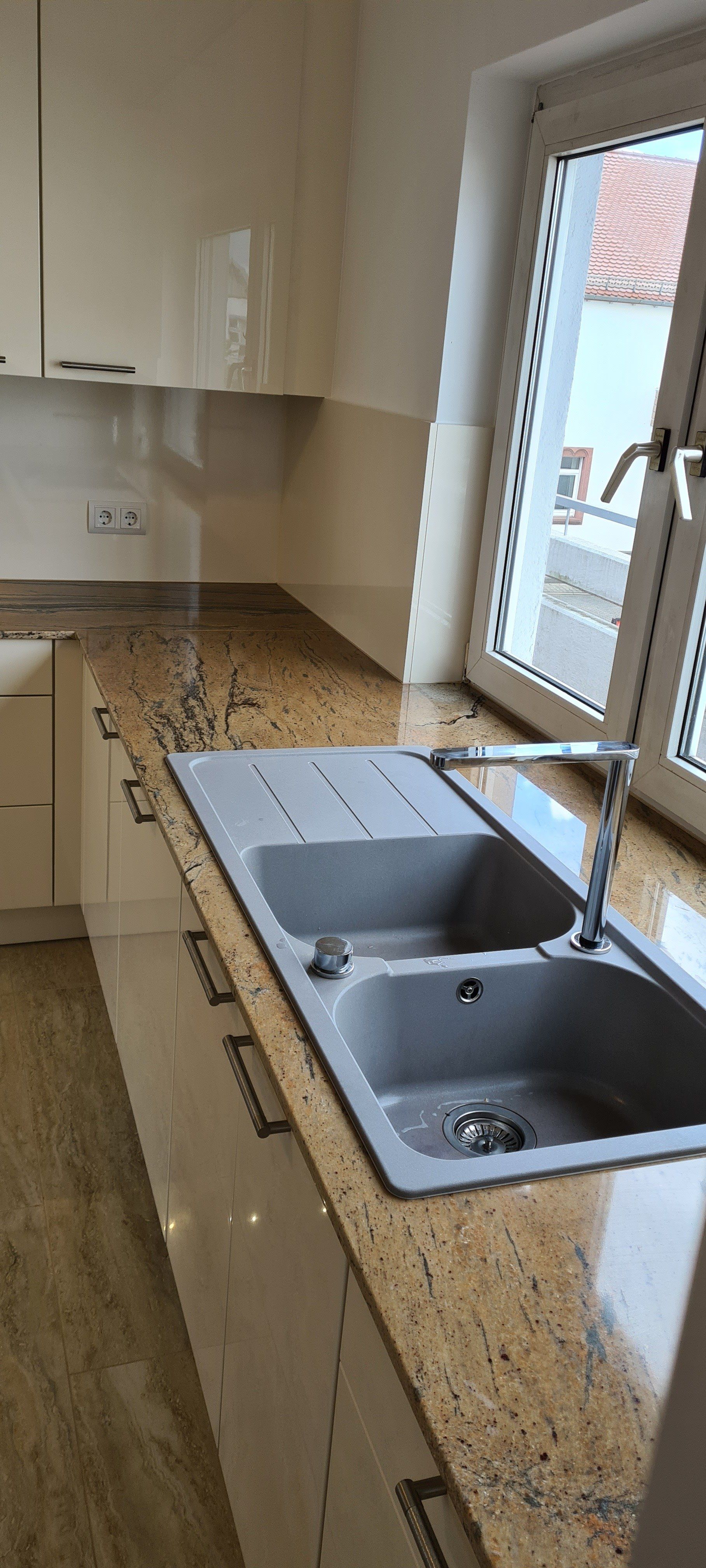 Verleihen Sie Ihrer Küche einen Hauch von Eleganz und Raffinesse mit einer polierten Natursteinarbeitsplatte in Praha Gold und glänzenden Magnolia Hochglanzfronten. Die beiden Becken mit Abtropffläche und die innovative Vor-Fenster-Armatur von Blanco setzen neue Maßstäbe für modernes Küchendesign und Funktionalität.