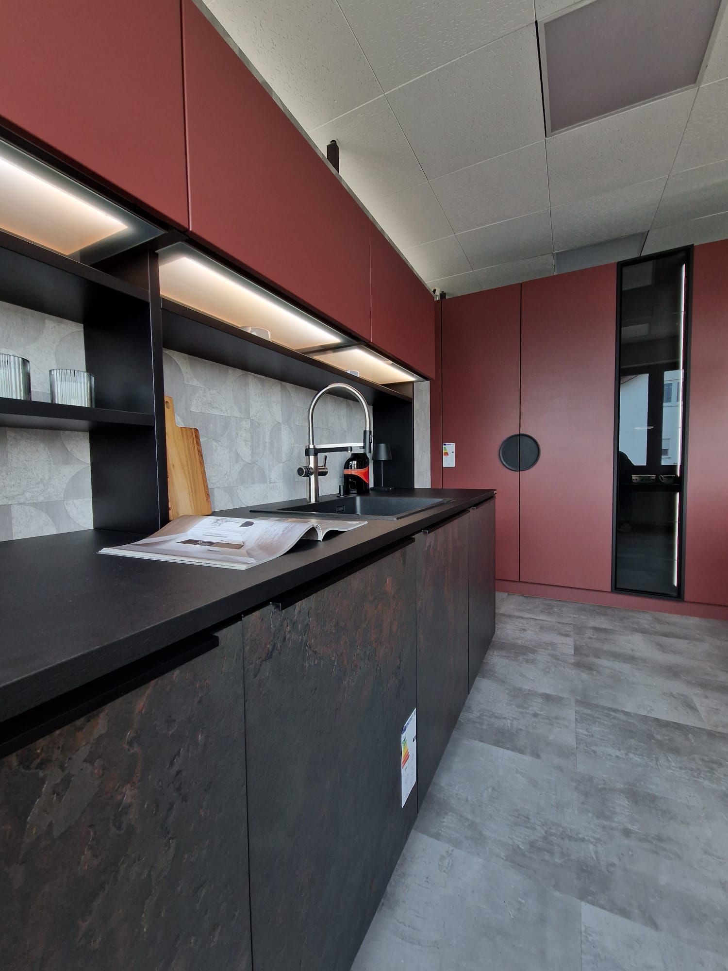 Unsere Nolte Neo Einbau-Musterküche ist das ultimative Symbol für modernen Luxus, mit Features wie der Blanco Drink.hot Evol-S Pro für jederzeit verfügbares, heißes Wasser, sowie Fronten in Schiefer-Schwarz Rosato und Henarot Softlack, einer Berbel Skyline Dunstabzugshaube und beleuchteten Hängeschränken mit elektrischer Öffnung.