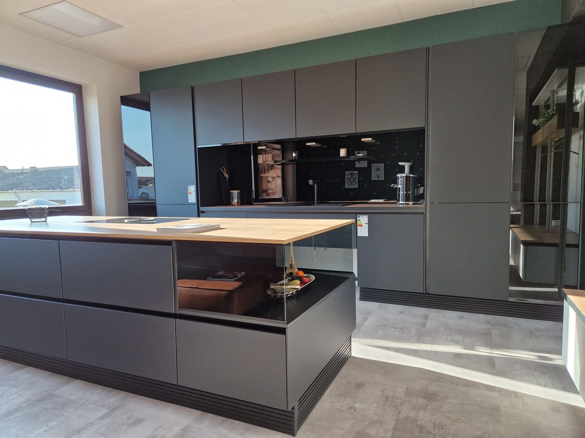 Entdecken Sie die Faszination einer ganz schwarzen Einbau-Musterküche ohne Griffe, die mit einer Keramik-Arbeitsplatte in Holzoptik und einer eleganten Vitrine für Gläser Ihre Küche in ein modernes Meisterwerk verwandelt.