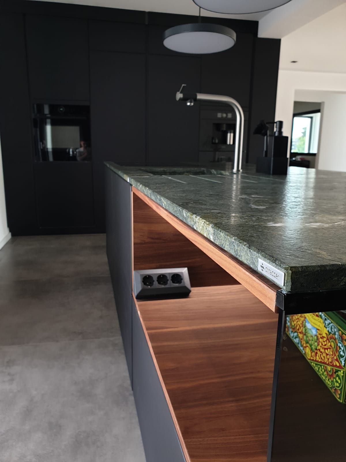 Machen Sie Ihre Küche zum Helden des Raums mit unserer Nobilia Einbauküche, die mit Antifingerprint Fronten in Schwarz und einer Natursteinarbeitsplatte von Strasser in Alpengrün im Leather-Look die Essenz von Stil verkörpert, während die elektrisch öffnende Insel von GRASS und die Einbaugeräte von Bosch AccentLine die Funktionalität auf ein neues Niveau heben.