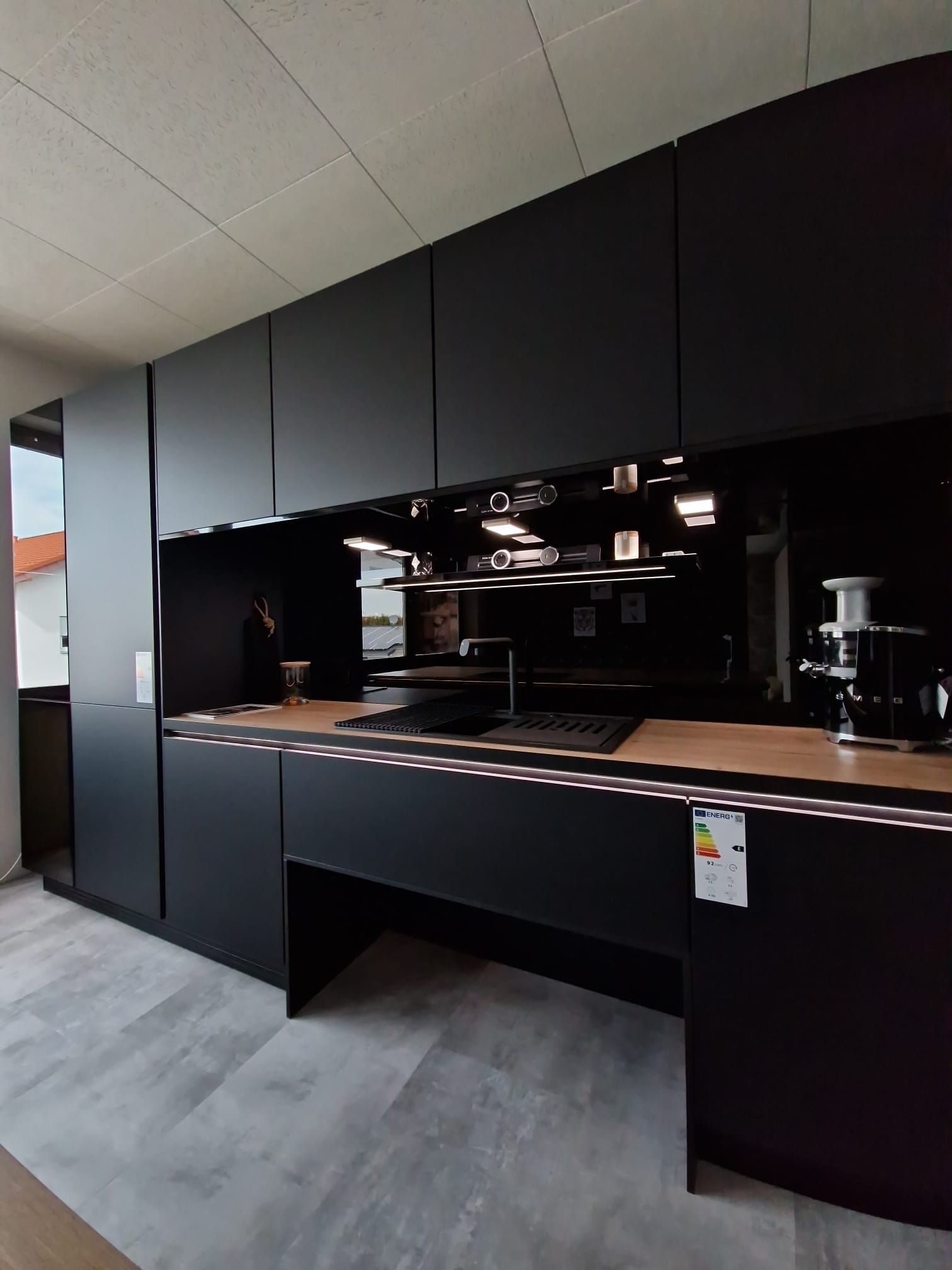 Mit unserer Einbau-Musterküche in Schwarz holen Sie sich ein Stück Luxus in Ihr Zuhause. Die Keramik-Arbeitsplatte in Holzoptik und die elegante Vitrine für Gläser verleihen Ihrer Küche eine exklusive Note.