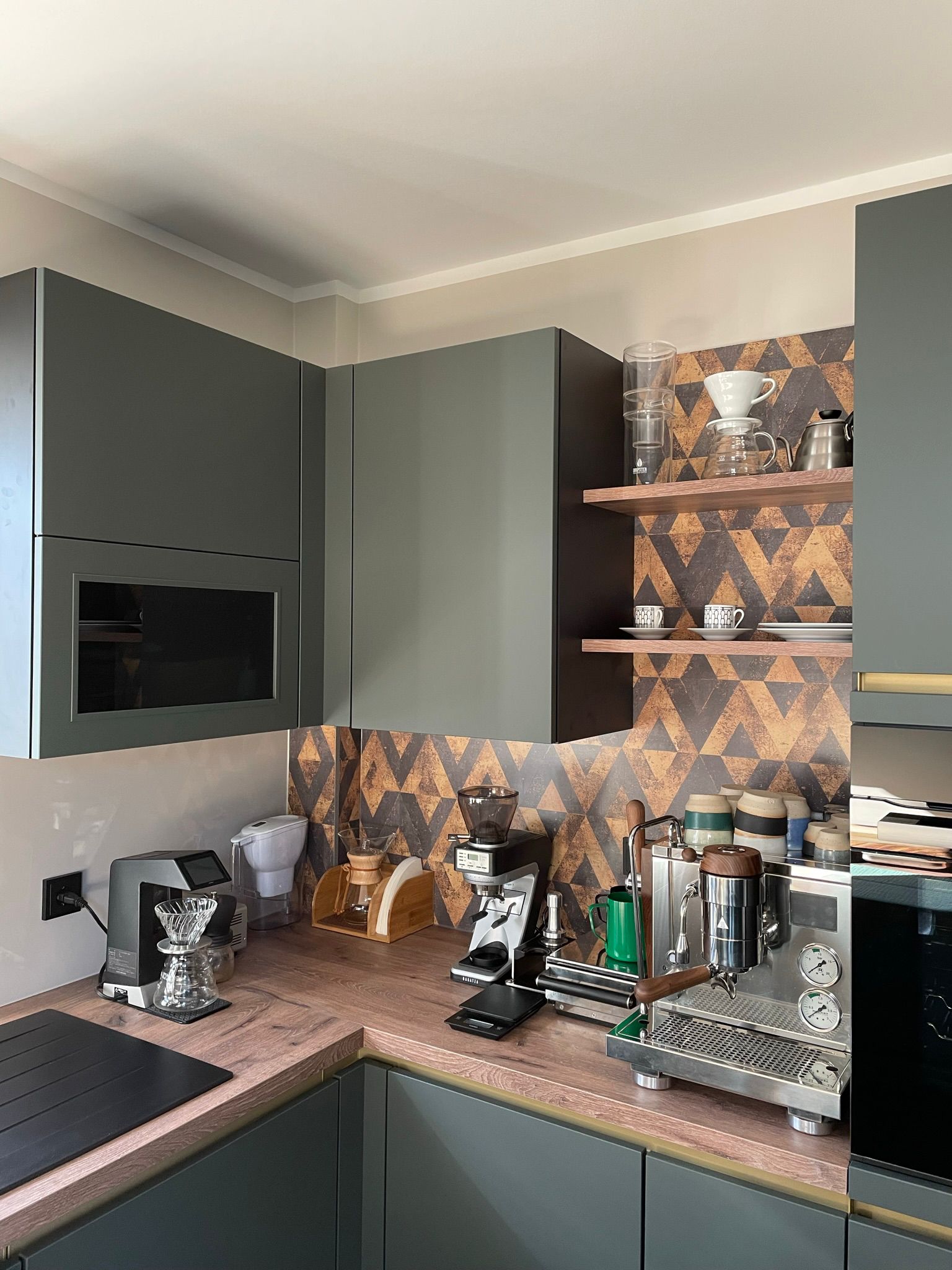 Auf dem Bild präsentiert sich eine ansprechende Nobilia Küche mit grifflosen Schränken, die durch ihre EASYTOUCH 964 Mineralgrün ultramatten Fronten einen modernen und edlen Touch erhält. In dieser Küchenecke, die wie eine Oase gestaltet wurde, kann man sich morgens herrlich bei einer Tasse Kaffee entspannen. Die sorgfältig ausgewählten Bosch accentline Einbaugeräte in Schwarz fügen sich nahtlos in das Design ein und unterstreichen die harmonische Farbgebung der Mineralgrünfronten. Diese Küche strahlt eine Atmosphäre der Ruhe und des Genusses aus, perfekt für entspannte Kaffeemomente am Morgen.