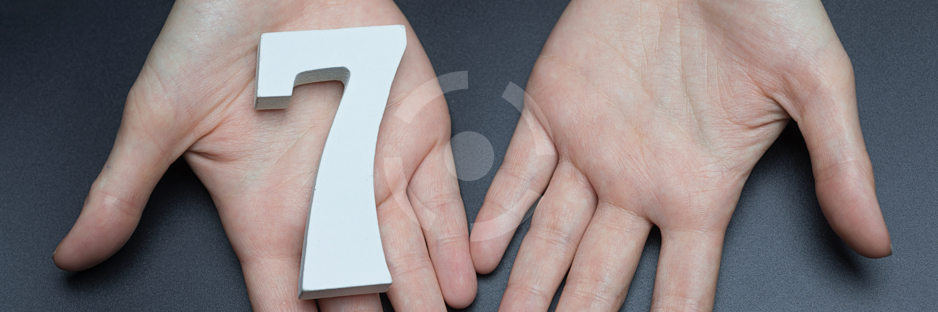Auf dunklem Hintergrund befindet sich die Zahl 7 auf einer Handfläche von zwei dargestellten Händen