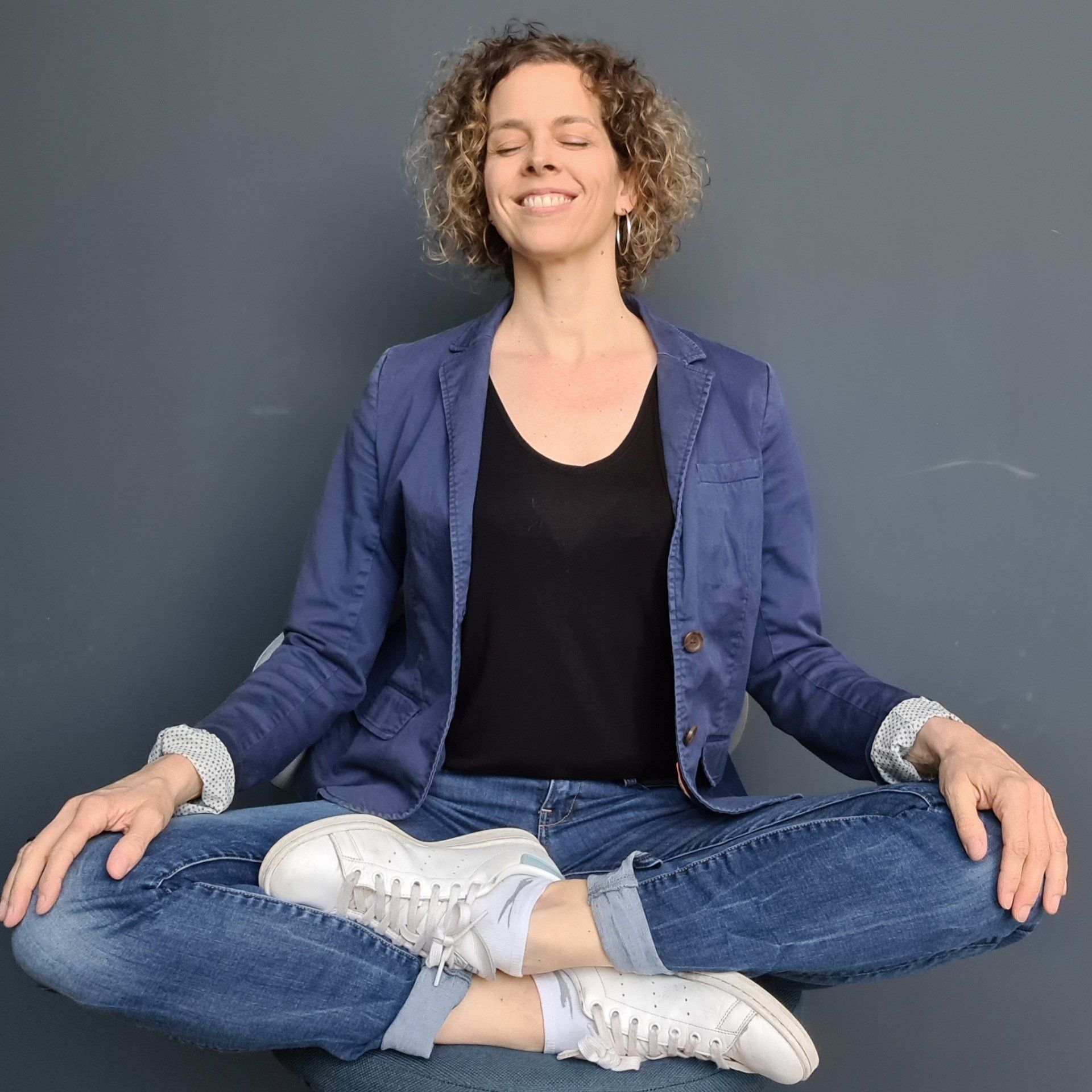 Kerstin Markovic sitzt im Schneidersitz mit geschlossenen Augen auf einem Stuhl und übt Achtsamkeit