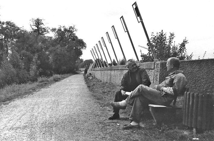Zwei Männer auf Bank sitzend, Kiesweg, Rasen, Mauer mit Metallstelen