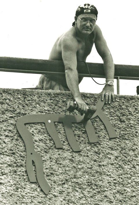Mann mit nacktem Oberkörper schraubt mit einem Akkuschrauber ein Metallstück an eine Mauer
