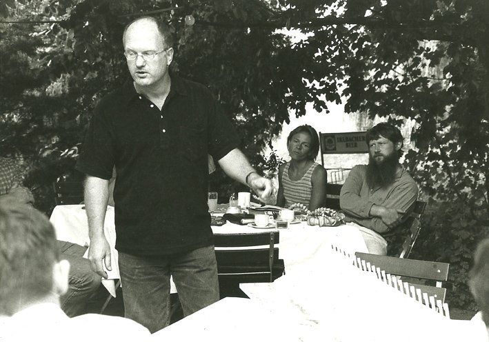 Stehender Mann in schwarzem T-Shirt. Im Hintergrund sitzend bärtigr Mann und Frau.