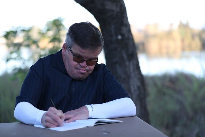 Kristofer, the Author, seated outdoors at a table writing, foliage and water in the background.