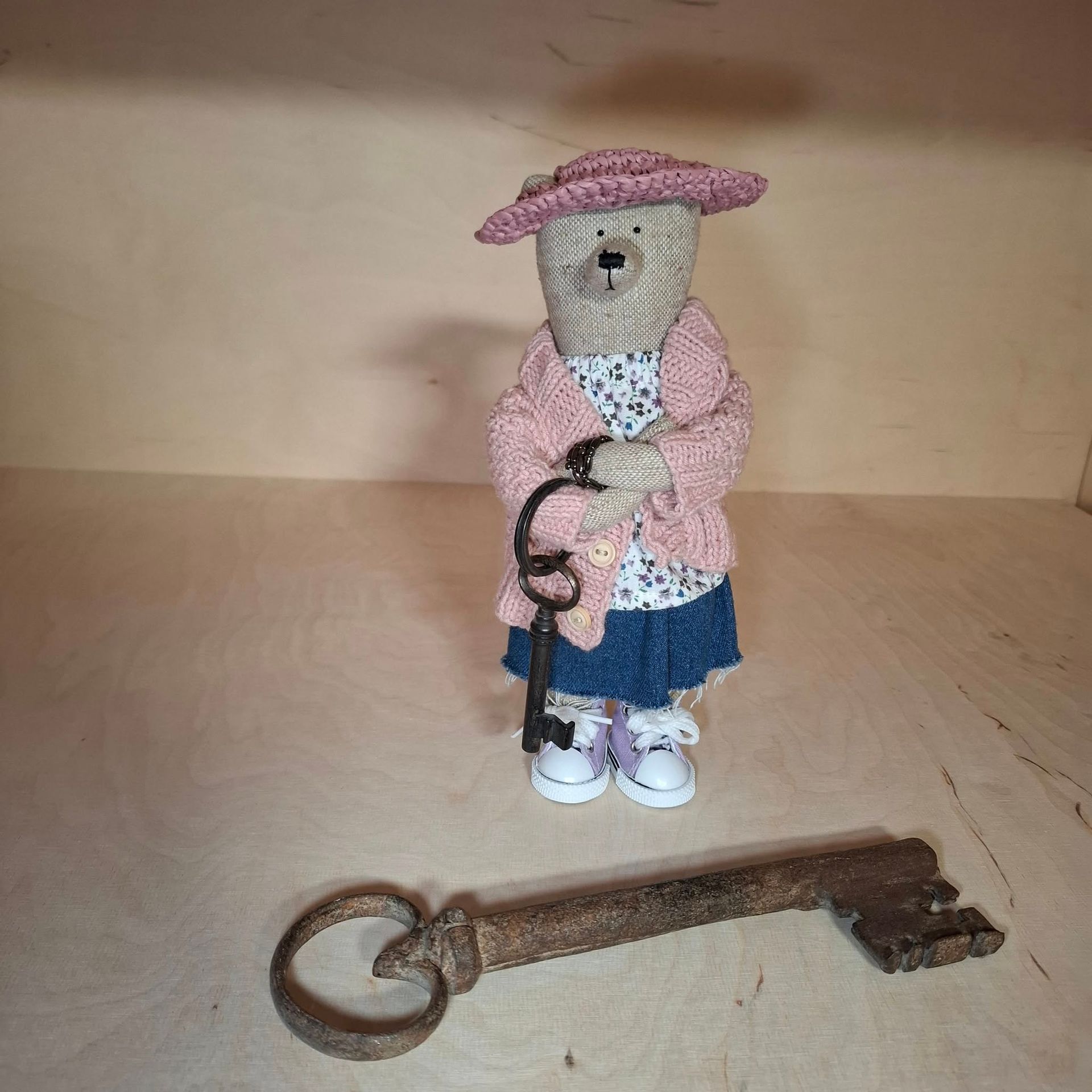 Bärin Freya steht mit einem Schlüsselbund in ihrem leeren Laden. Vor ihr liegt ein großer alter magischer Schlüssel. Der Teddybär trägt eine rosa Strickjacke, einen blauen Jeansrock und rosa Strohhut.