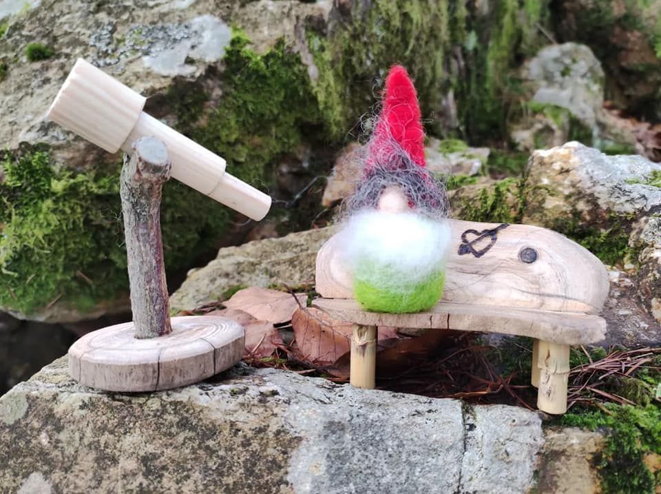 Ein grüner Wichtel mit roter Mütze und weißem Bart sitzt auf einem Felsen auf seiner Holzbank mit Herz und schaut in sein Fernrohr.