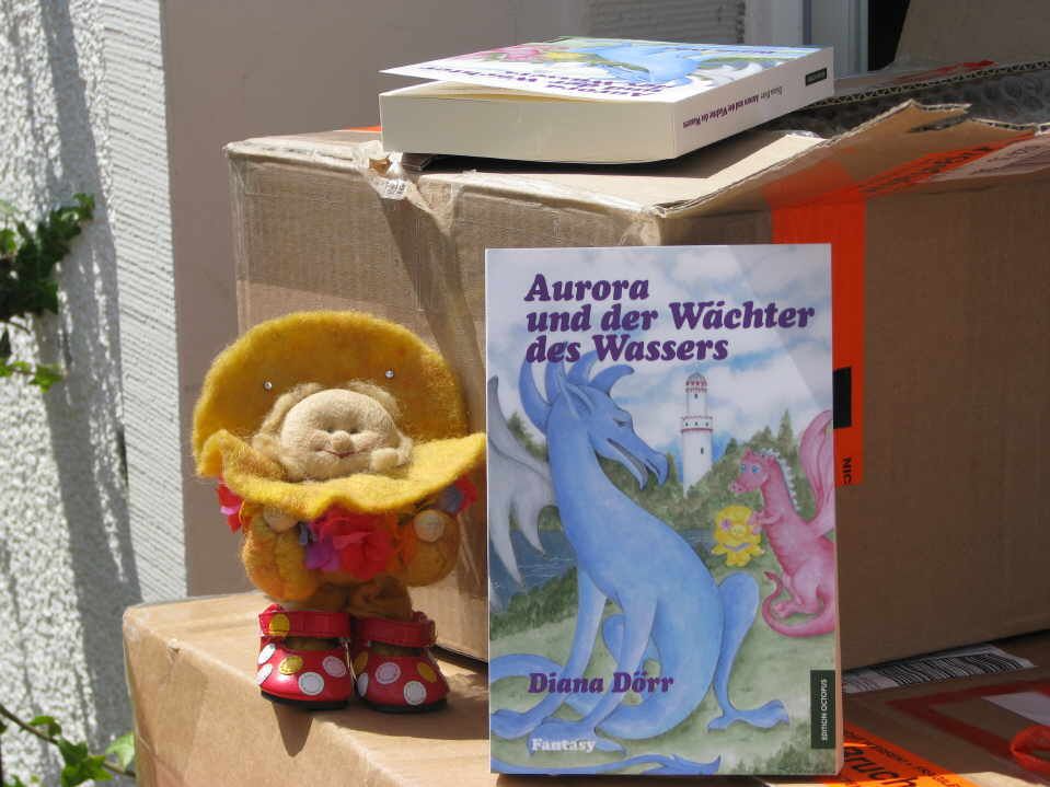 Der gelbe Sonnenengel Aurora steht stolz vor dem frisch gedruckten Buch mit dem Titel Aurora und der Wächter des Wassers
