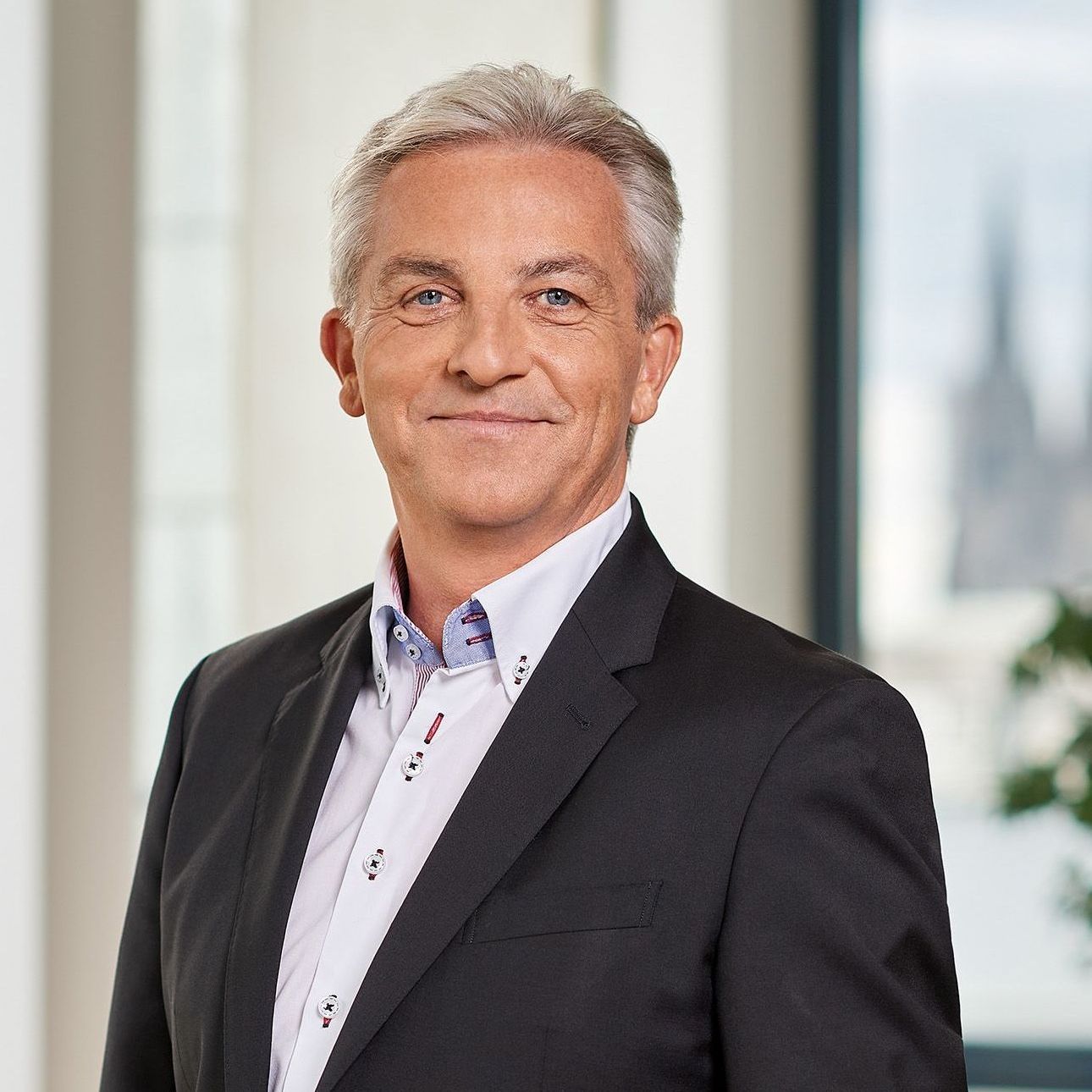 Ein lächelnder Mann mit grauen Haaren, der ein dunkles Sakko und ein weißes Hemd trägt. Er steht in einem hellen Raum mit großen Fenstern im Hintergrund und lächelt freundlich.