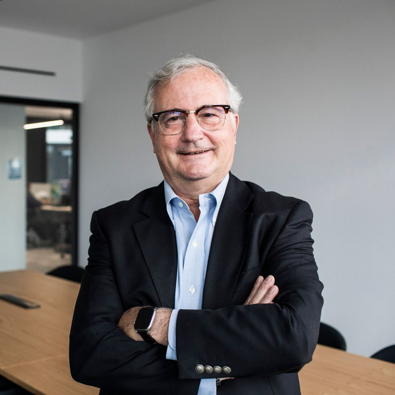 Ein lächelnder Mann mit Brille und weißen Haaren, der ein dunkles Sakko und ein weißes Hemd trägt. Er steht in einem großen Raum mit Tischen und hat die Arme vor der Brust verschränkt.