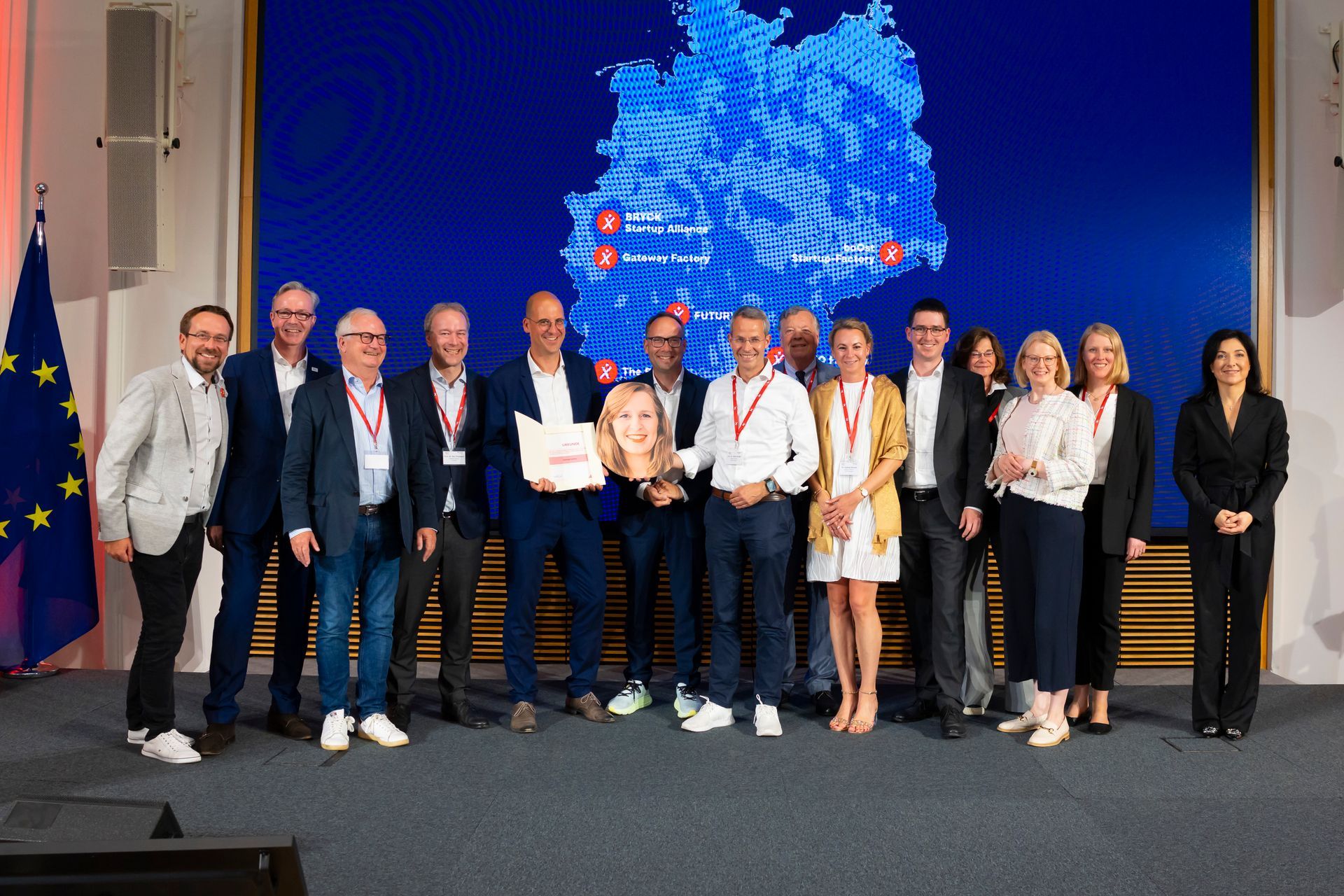 Eine Gruppe von fünfzehn Personen steht auf einer Bühne vor einer großen digitalen Karte Deutschlands. Eine Person hält eine Urkunde und alle lächeln in die Kamera. Links steht eine EU-Flagge.