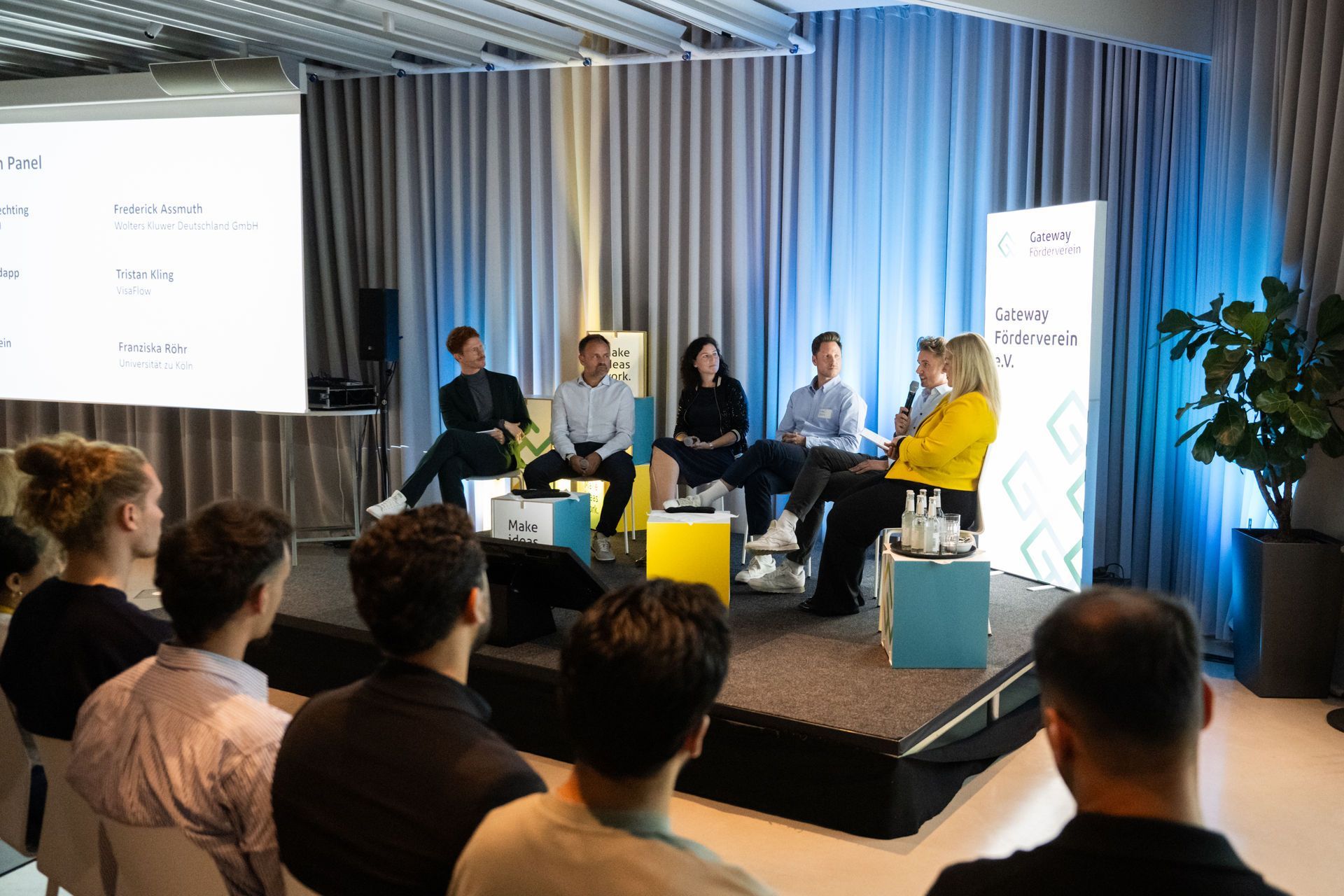 Mehrere Personen sitzen auf einer Bühne in einer Podiumsdiskussion. Eine Frau mit gelber Jacke spricht ins Mikrofon. Im Publikum sitzen Zuhörer, im Hintergrund ist eine Leinwand mit Namen und ein Banner des Gateway Fördervereins zu sehen.